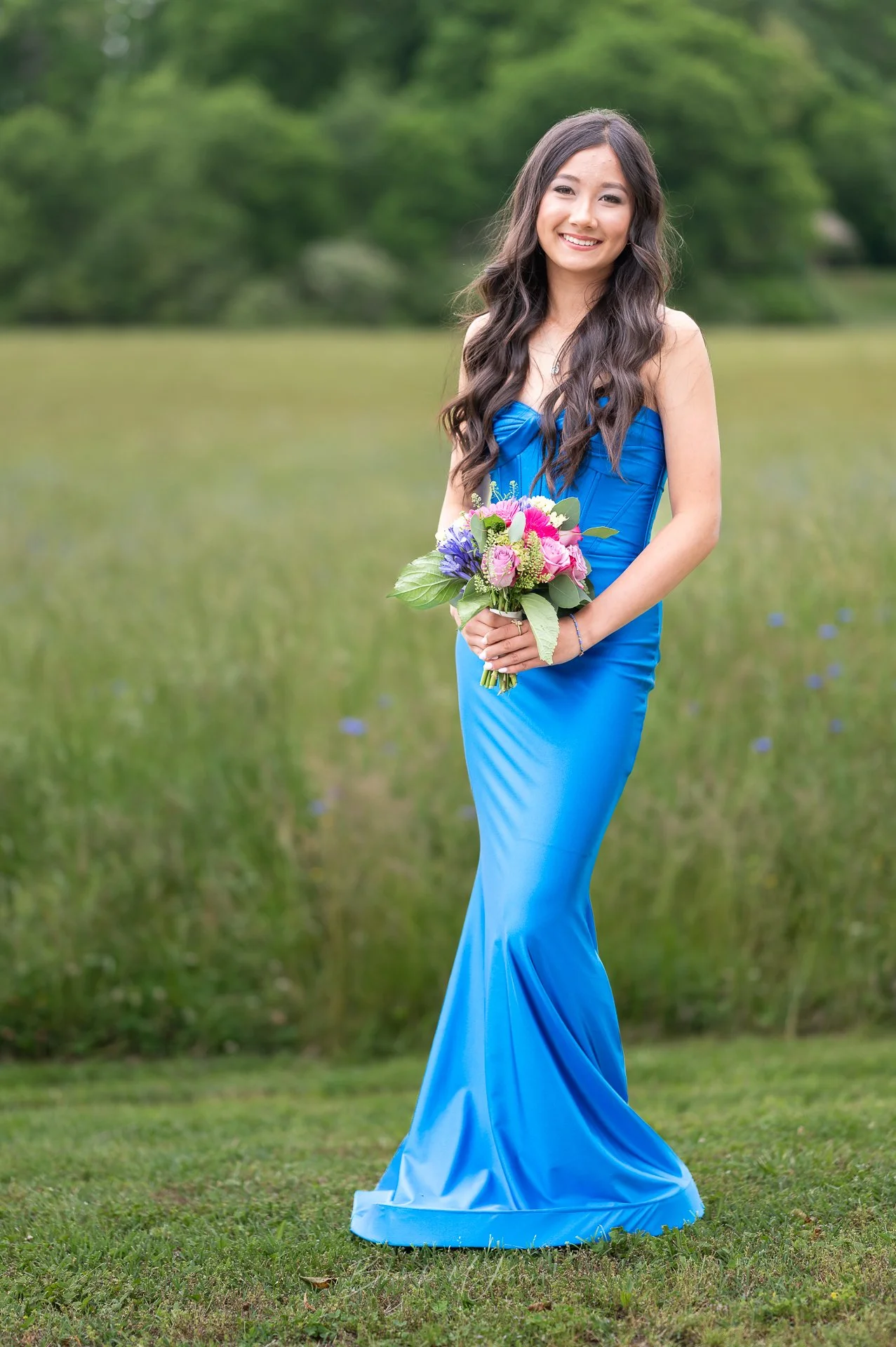 A young woman in a blue gown holding a colorful bouquet of flowers outdoors in a grassy area with trees in the background.