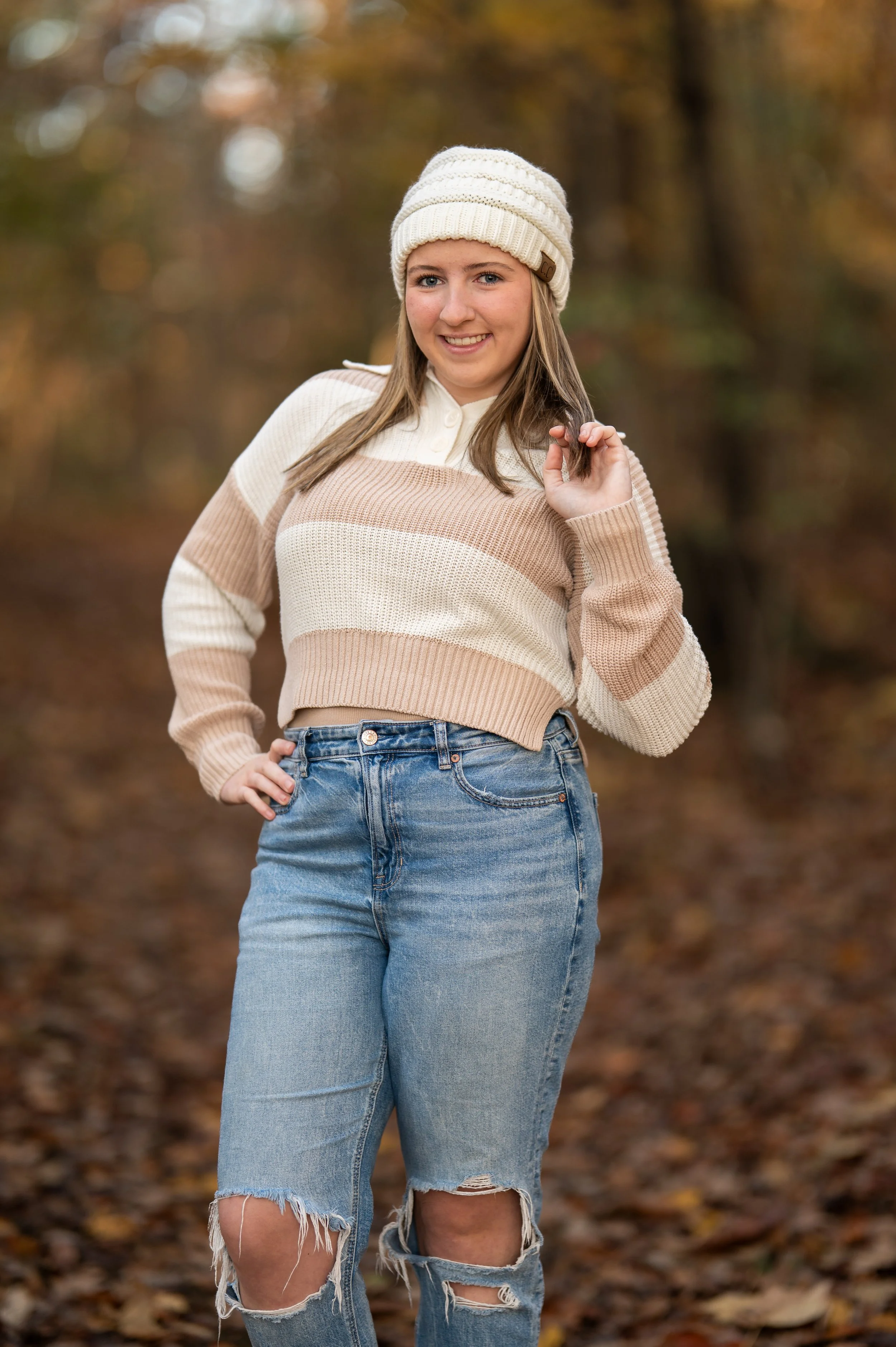 A young woman with long hair in a cream beanie hat, smiling, wearing a beige and cream striped sweater, and ripped jeans, standing outdoors on a fall day with autumn leaves on the ground and trees in the background.