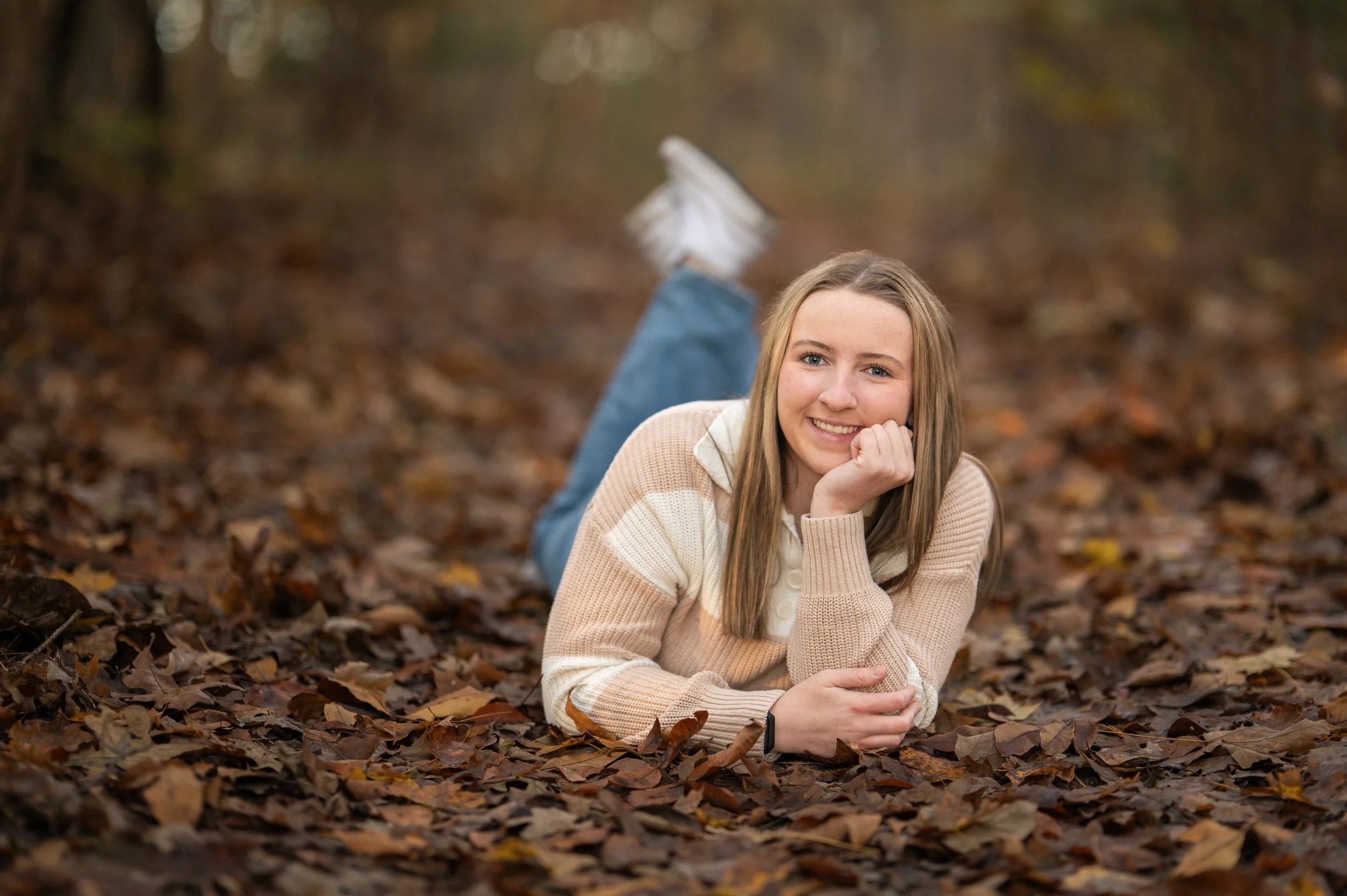 A young woman with long light brown hair, smiling, lies on her stomach on a bed of fallen autumn leaves in a forest, resting her chin on her hand.