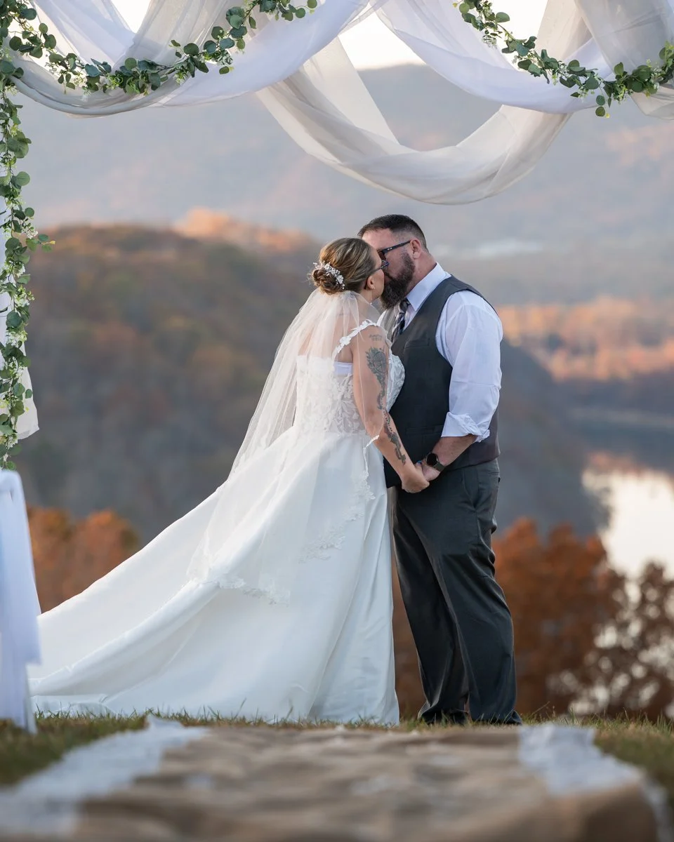 A bride and groom kissing under a decorated arch during an outdoor wedding, with mountains and trees in the background.