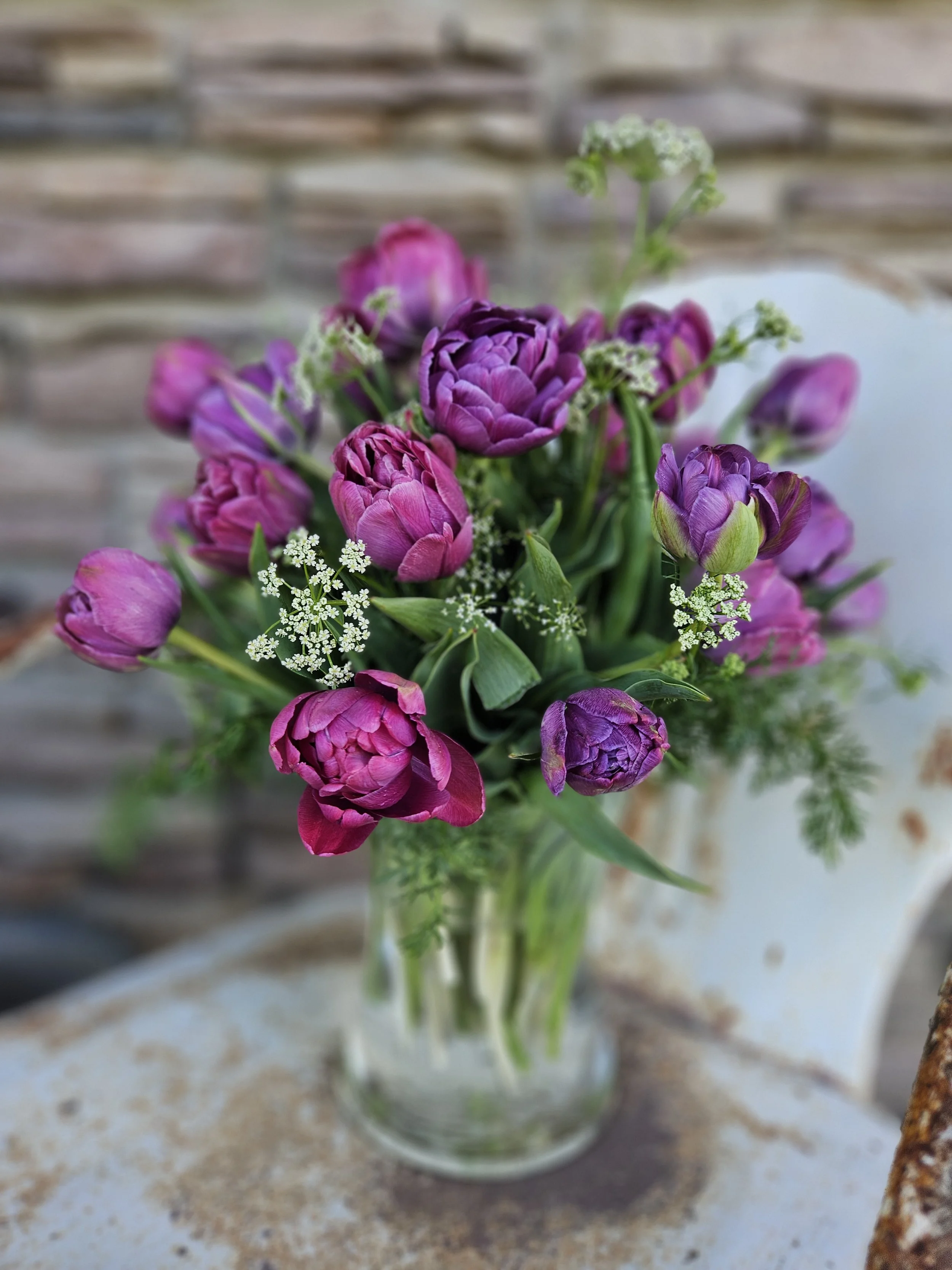 A vase of purple and pink tulips with small white flowers, placed on a weathered surface with a blurred brick wall background.