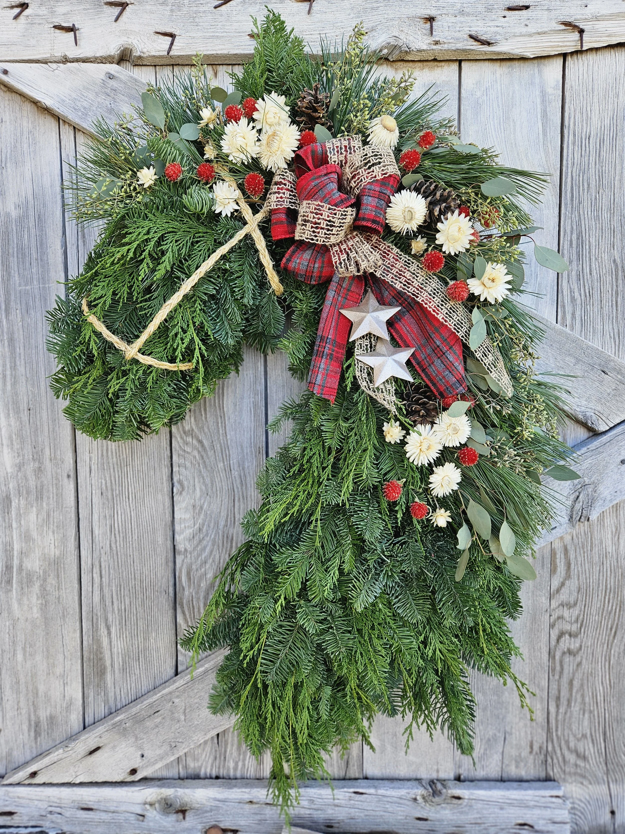 A Christmas wreath made of evergreen branches, decorated with red and white flowers, pinecones, a red plaid bow, silver stars, and a rope, hanging on a wooden door.