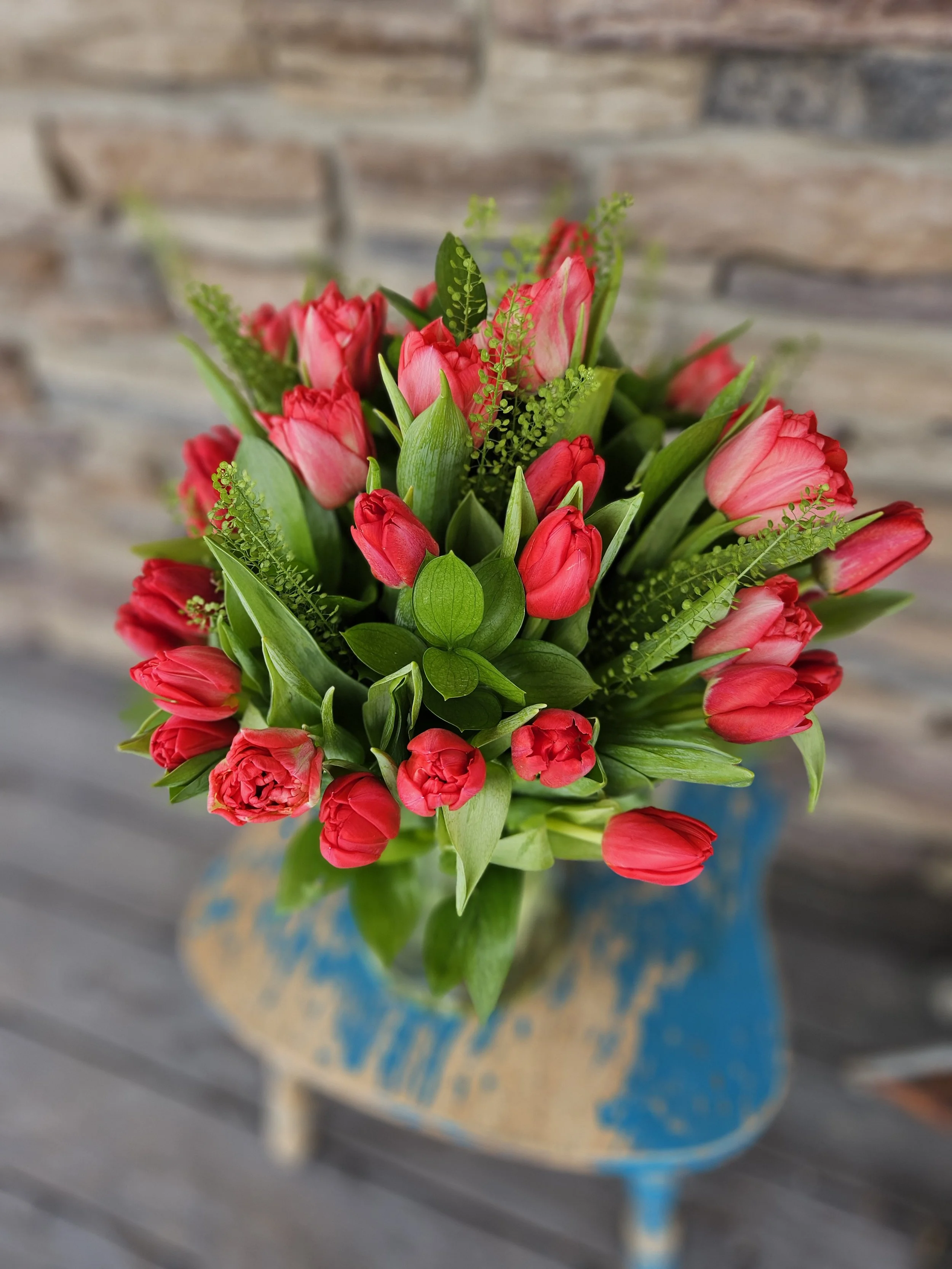A bouquet of lush red tulips, green foliage, in a glass vase on a weathered blue chairs with a wood and brick background.