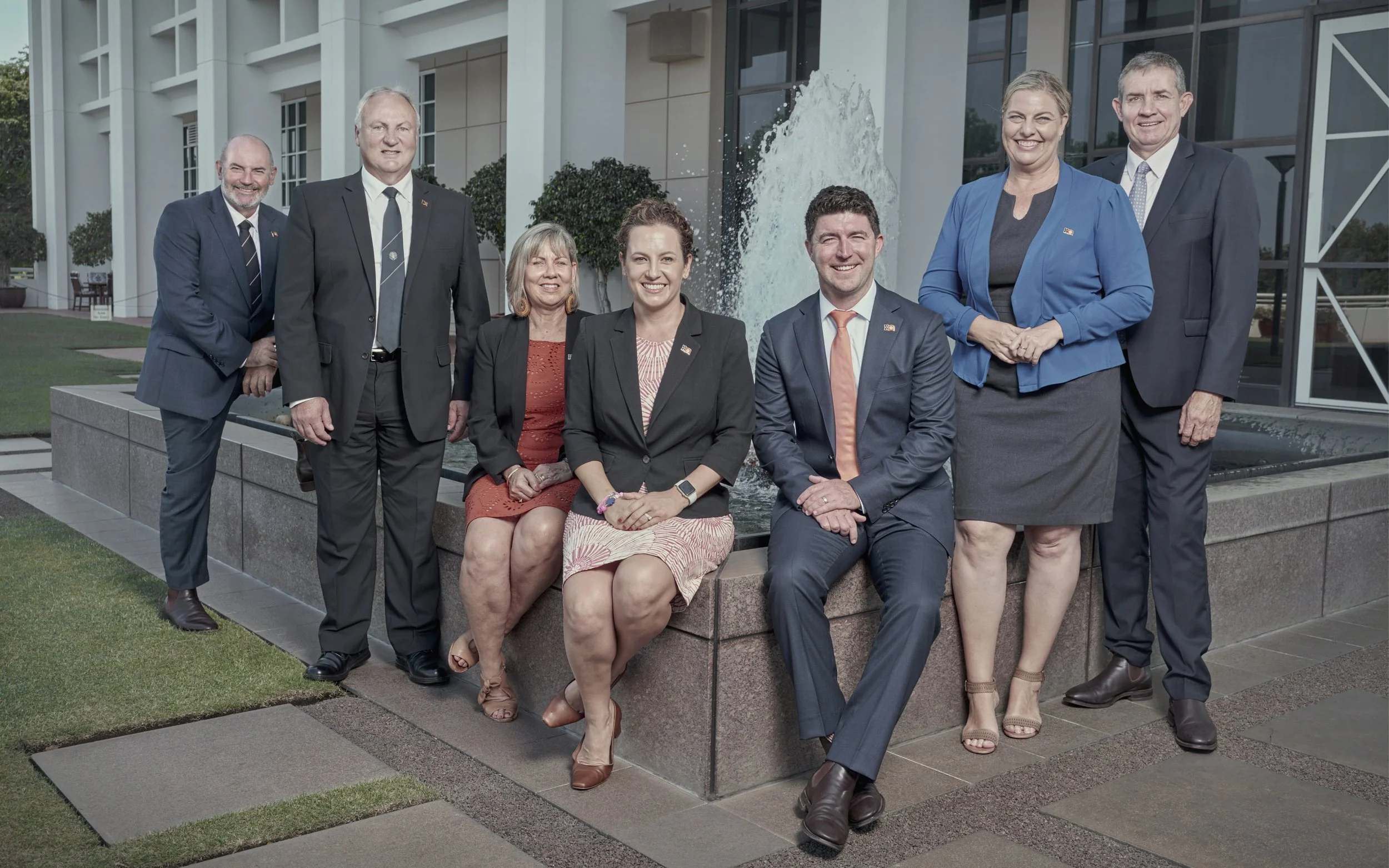 A group of nine professionally dressed people posing in front of a fountain outside a modern building.
