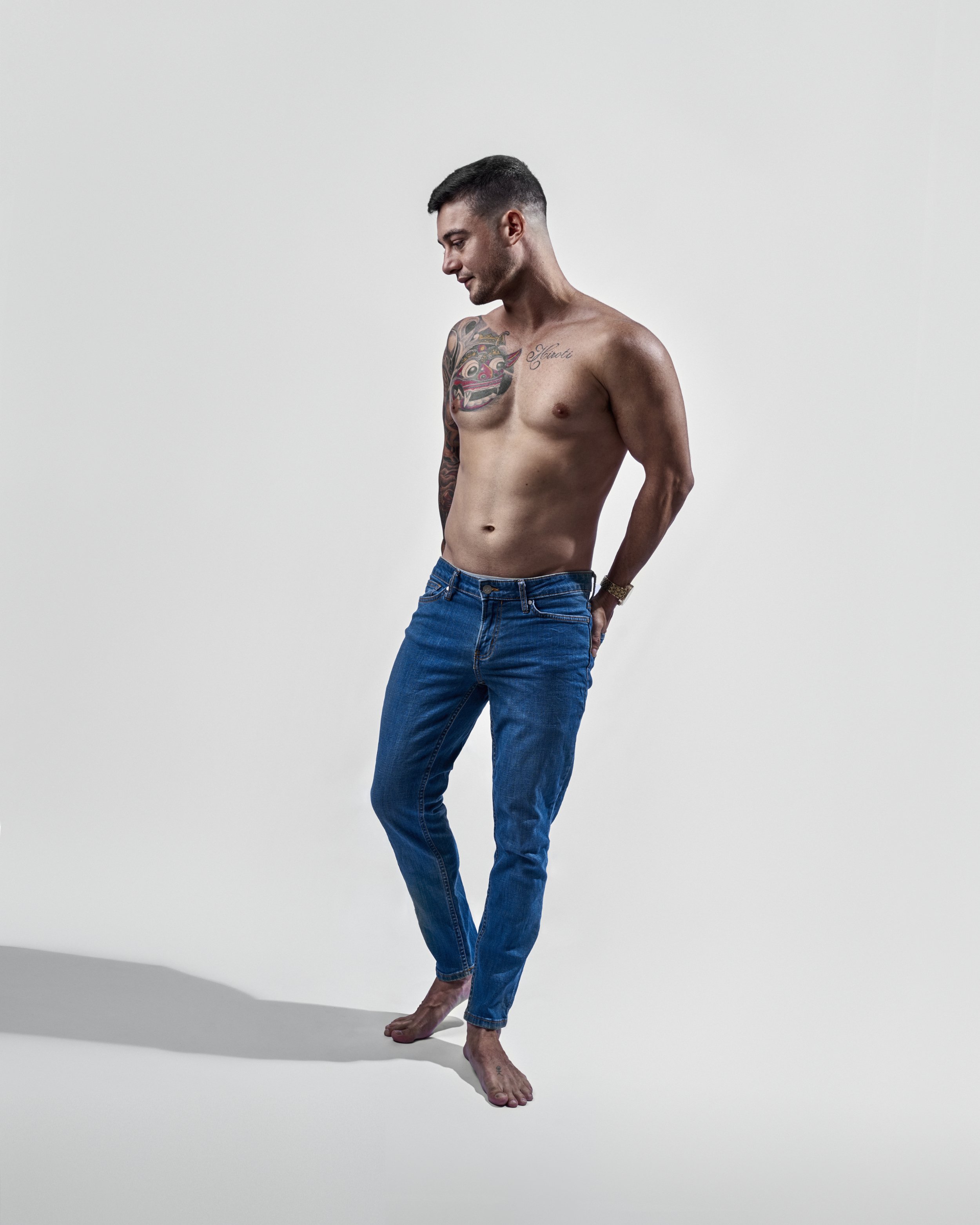 A shirtless young man with tattoos on his left shoulder and chest, standing on a white surface with a plain background, looking downward, wearing blue jeans.