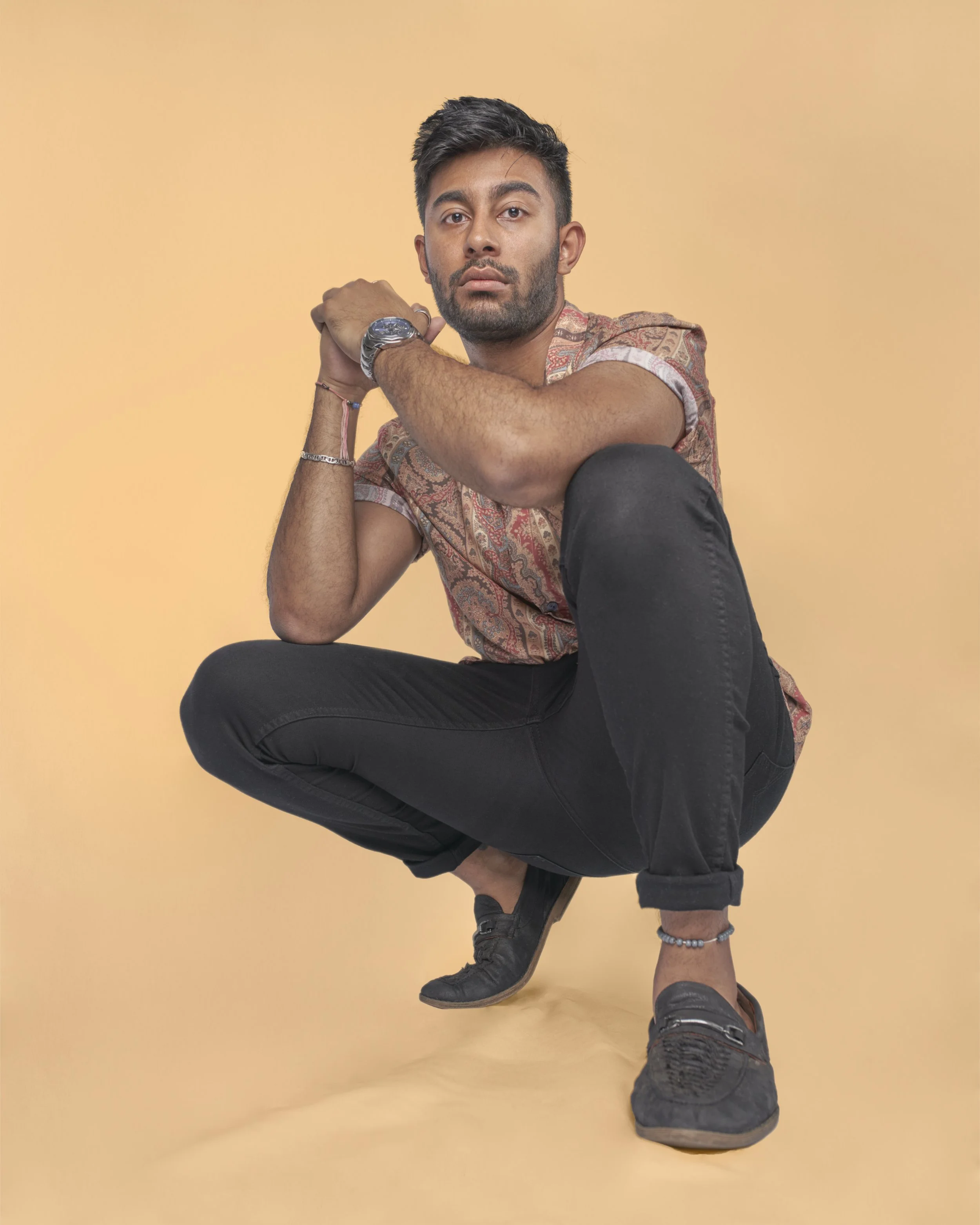 Young man crouching with one knee on the ground, wearing a patterned short-sleeve shirt, black pants, and black shoes, posing against a light orange background.