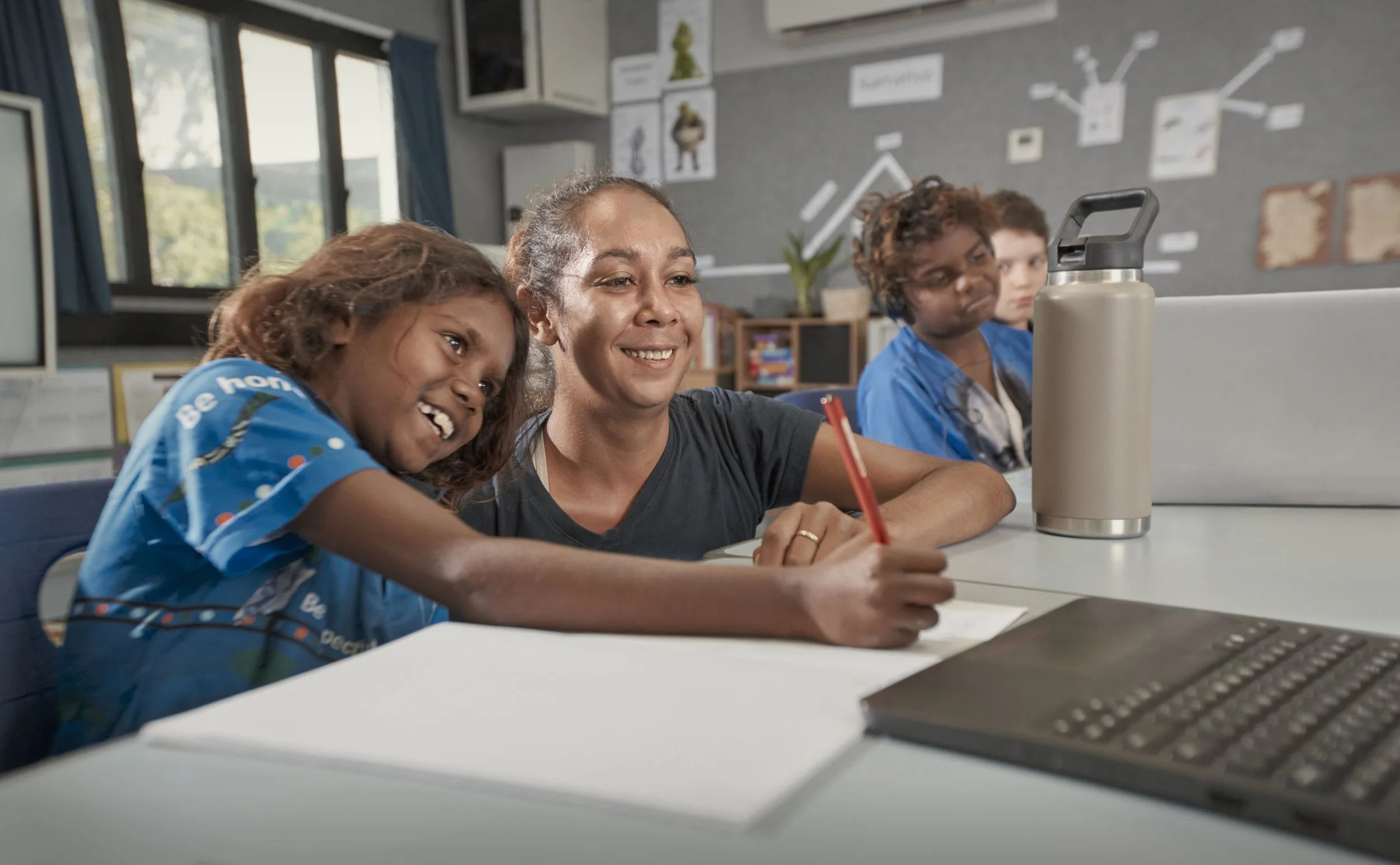 A teacher and two students in a classroom looking at a laptop screen, smiling with notebooks and a water bottle on the desk.