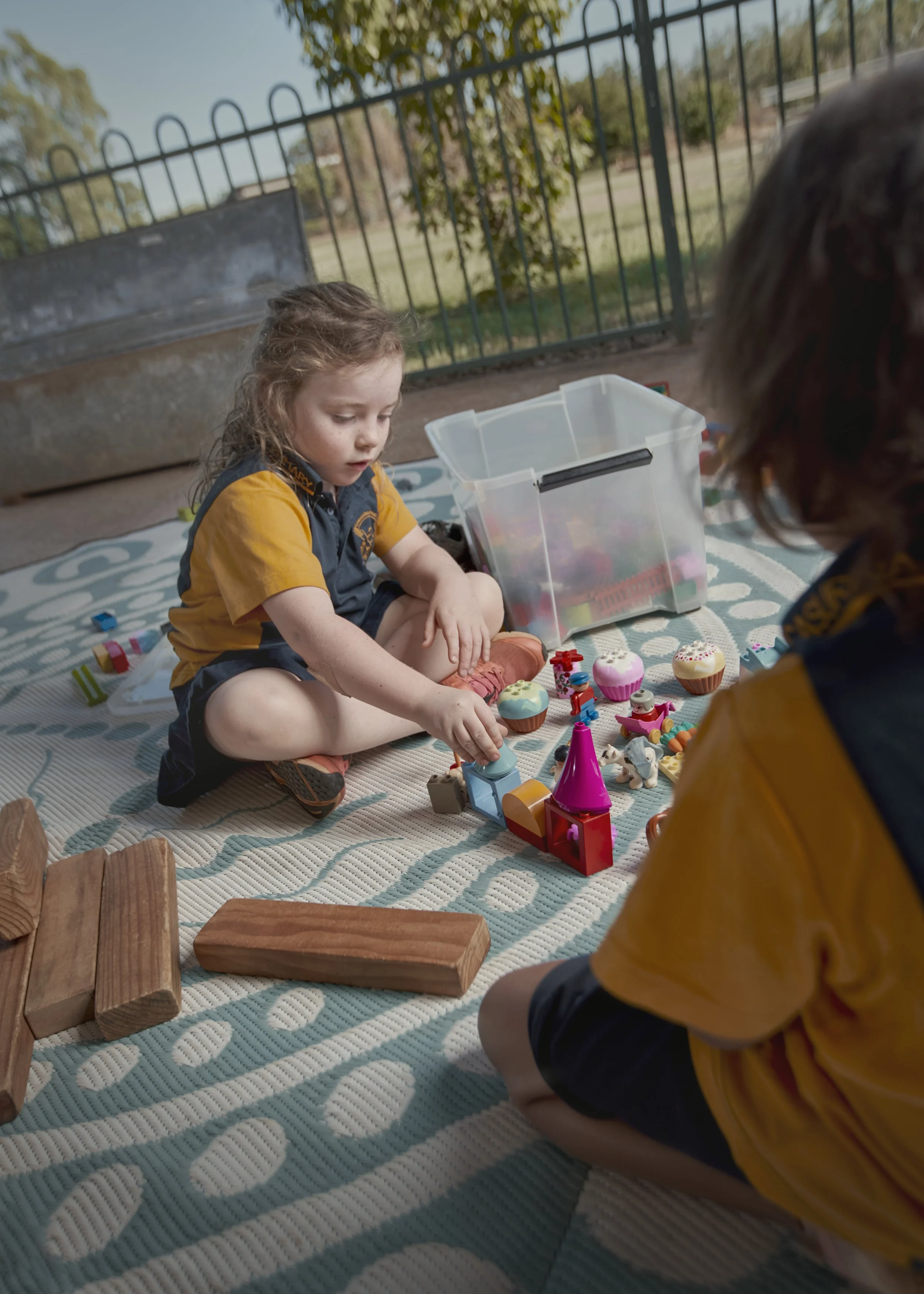 Two children playing with toys and building blocks on a blanket outdoors near a fence.