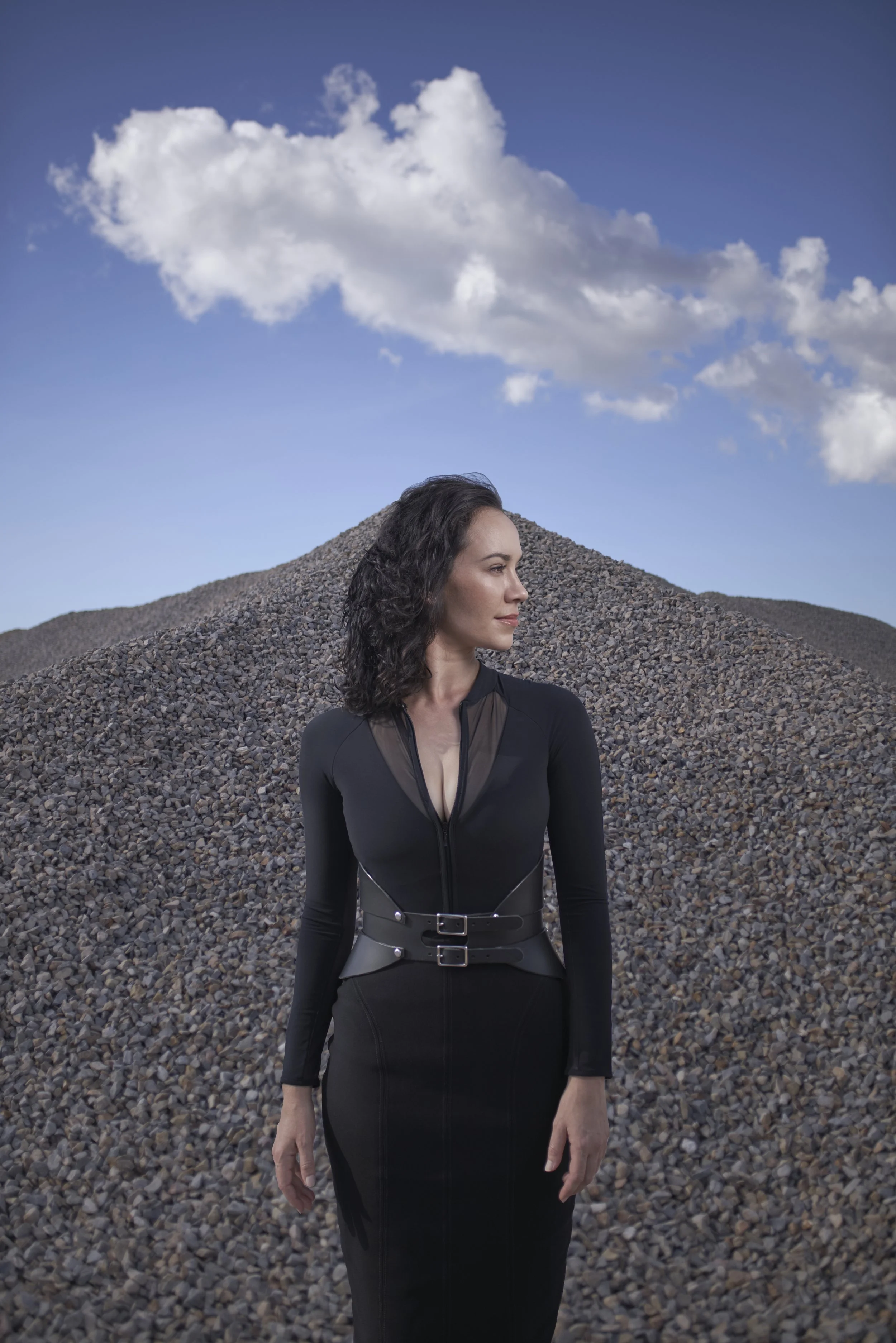 A woman stands in front of a large pile of small rocks or gravel, wearing a fitted black dress with sheer panels and a waist belt, with a blue sky and a large white cloud in the background.
