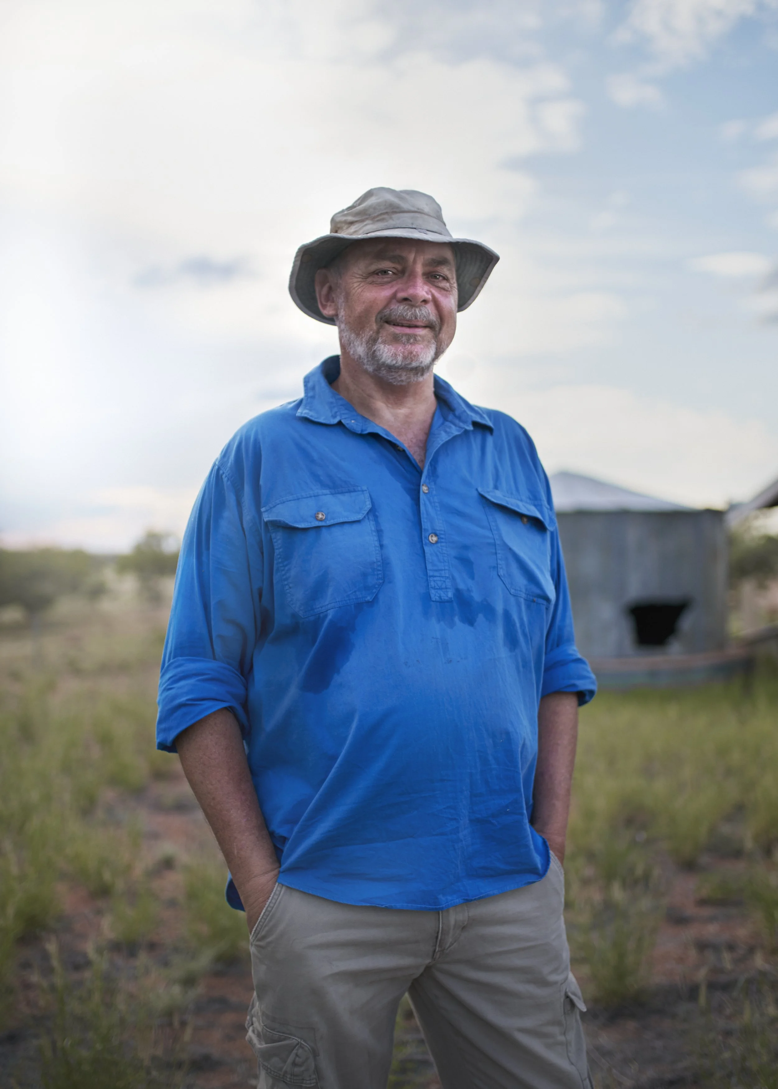 A man in a blue shirt and beige pants stands outdoors in a field, wearing a wide-brimmed hat, with a cloudy sky and a small barn in the background.