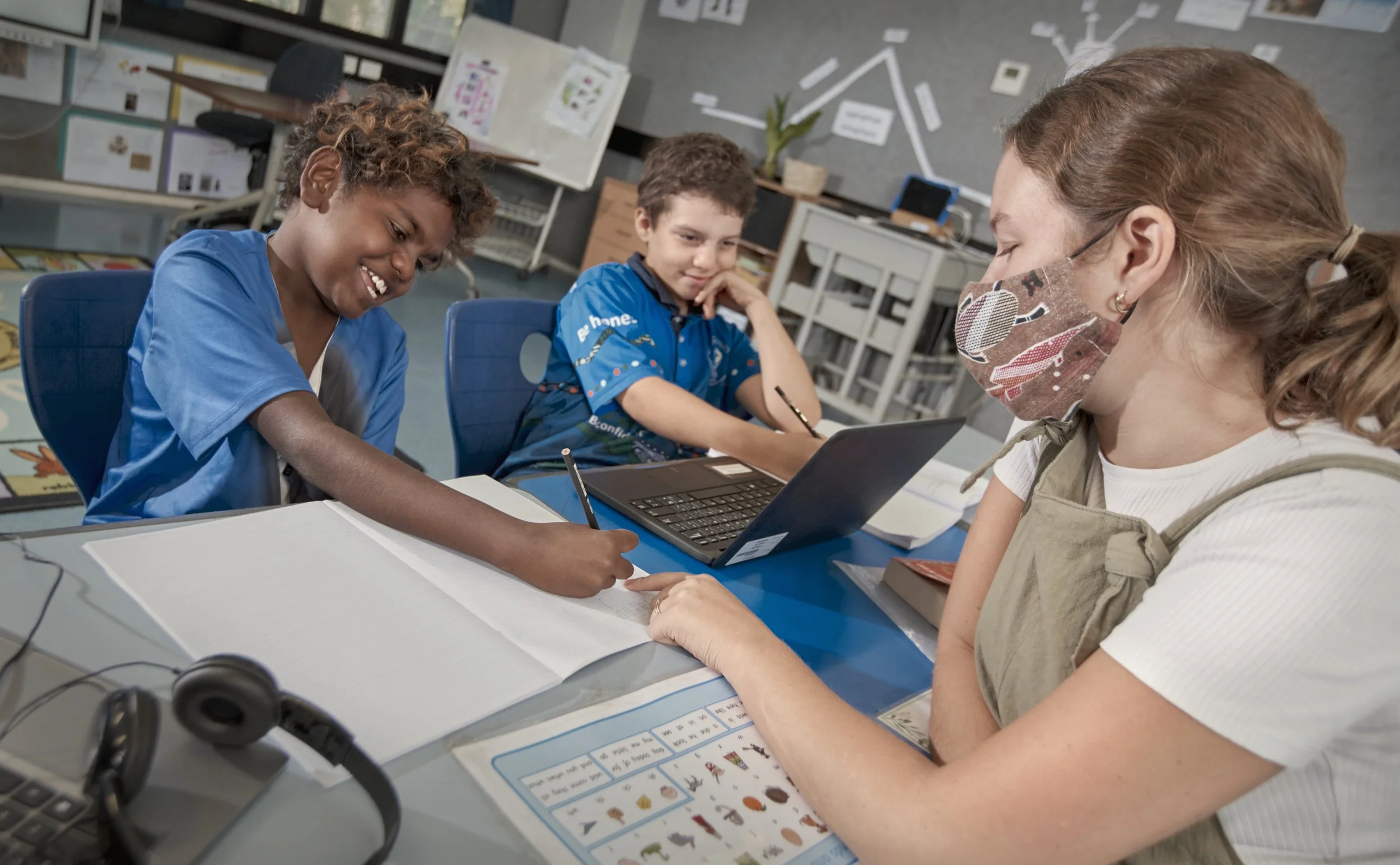 A teacher wearing a floral mask helps two young students with their work in a classroom. One student is smiling and writing in a notebook, while the other looks on thoughtfully.