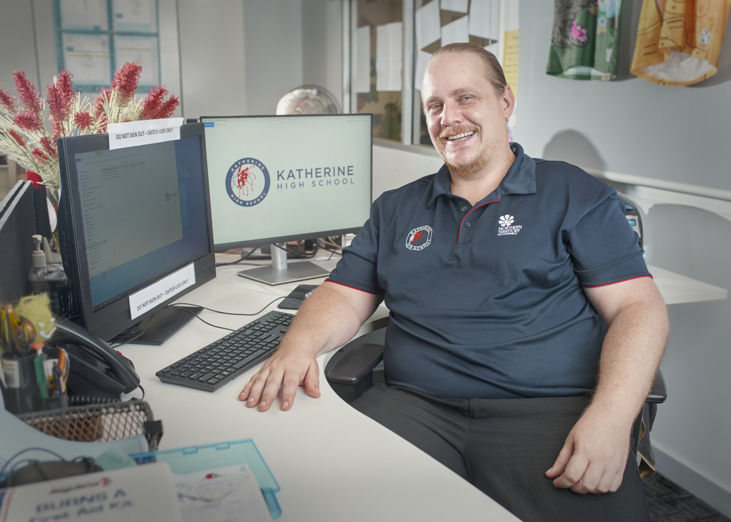A smiling man sitting at a desk with two computer monitors, one displaying a logo for Katherine High School, in an office environment.