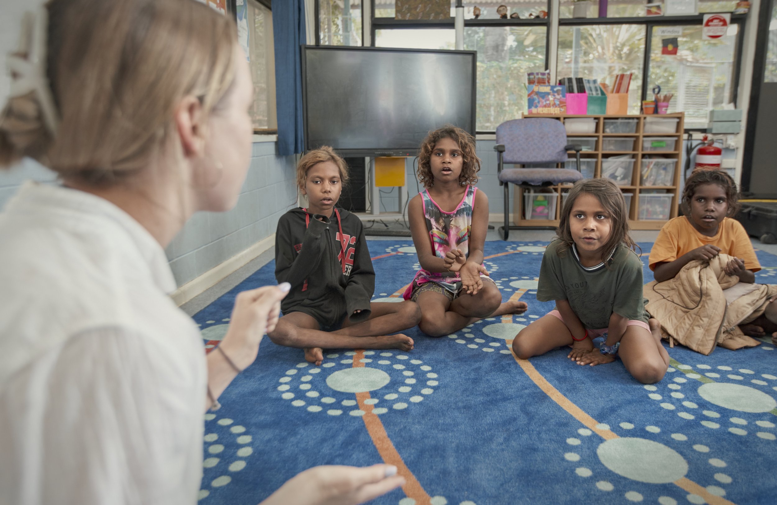 A woman appears to be talking to four young girls sitting on a colorful carpet in a classroom. The girls are attentively listening, with some looking surprised or engaged.