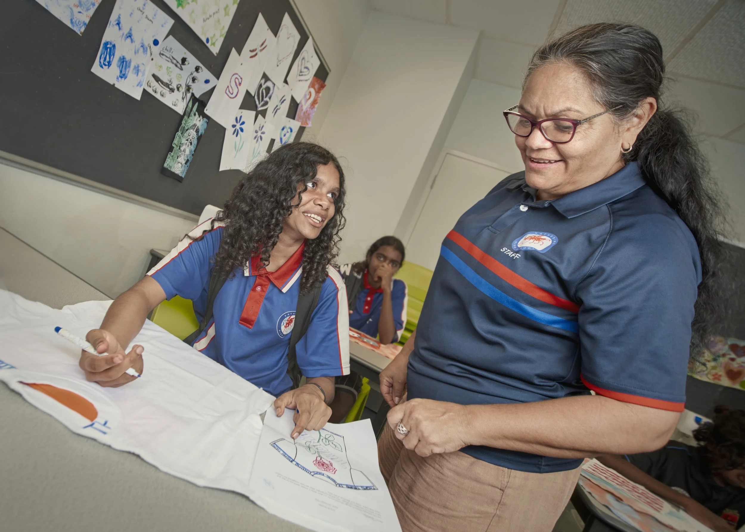 A young girl at a school desk converses with an older woman, both wearing blue uniforms with red and blue stripes, in a classroom with student artwork on the wall.
