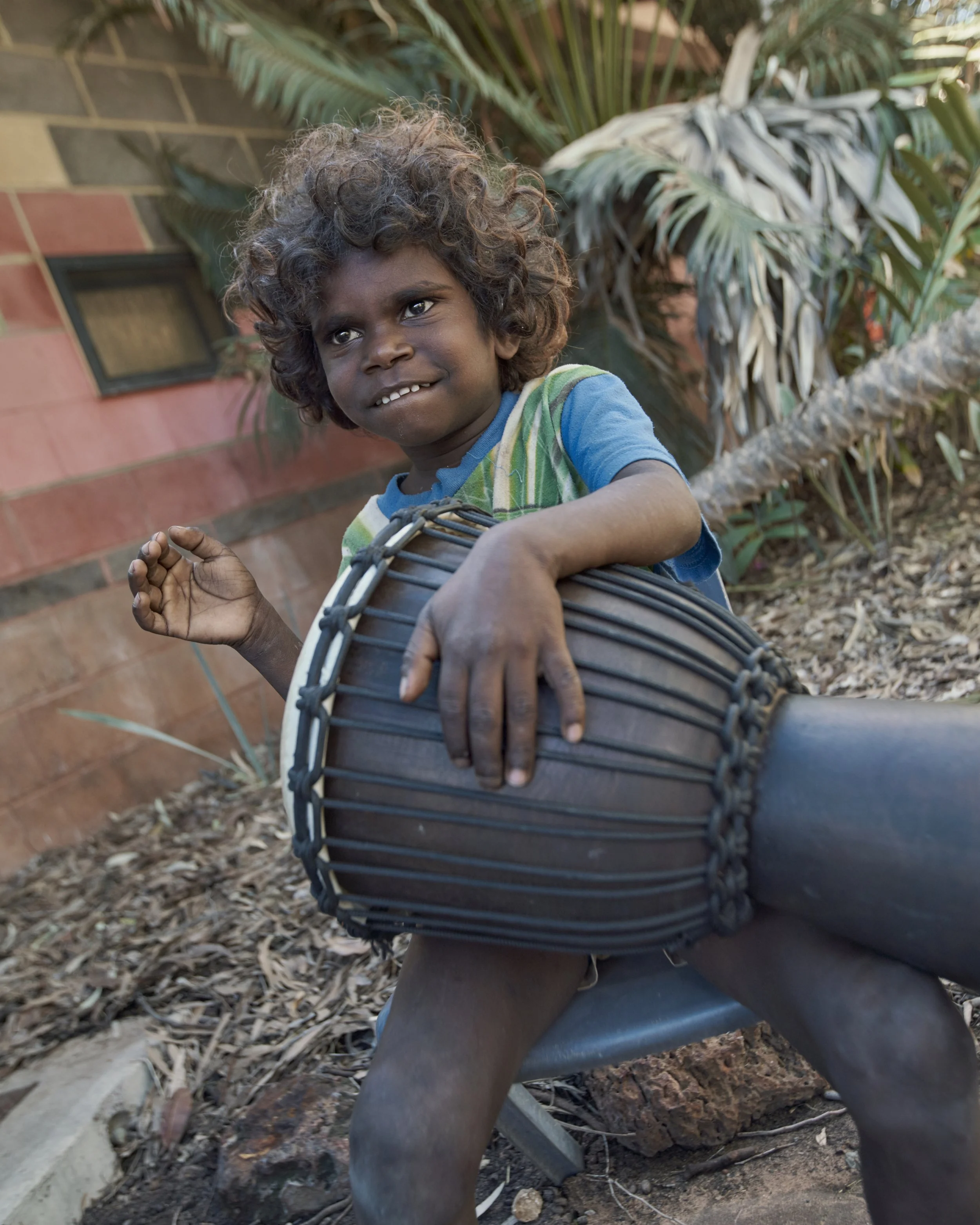 A young boy with curly hair playing a drum outdoors, with palm trees and a brick house in the background.