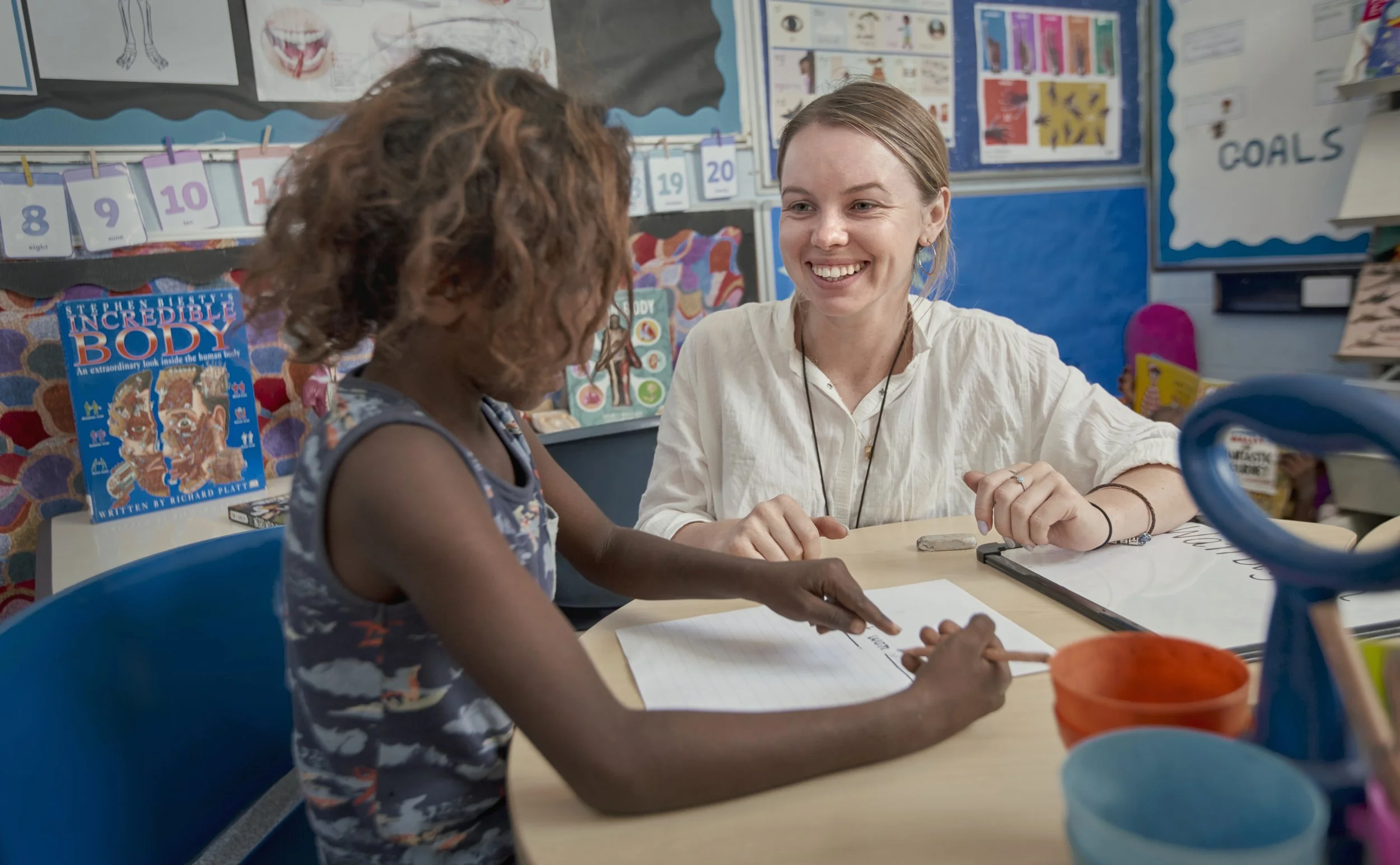 A teacher smiling at a young girl with curly hair in a classroom. The girl is writing on a worksheet. The classroom has educational posters, including one about the human body, and colorful supplies on the table.