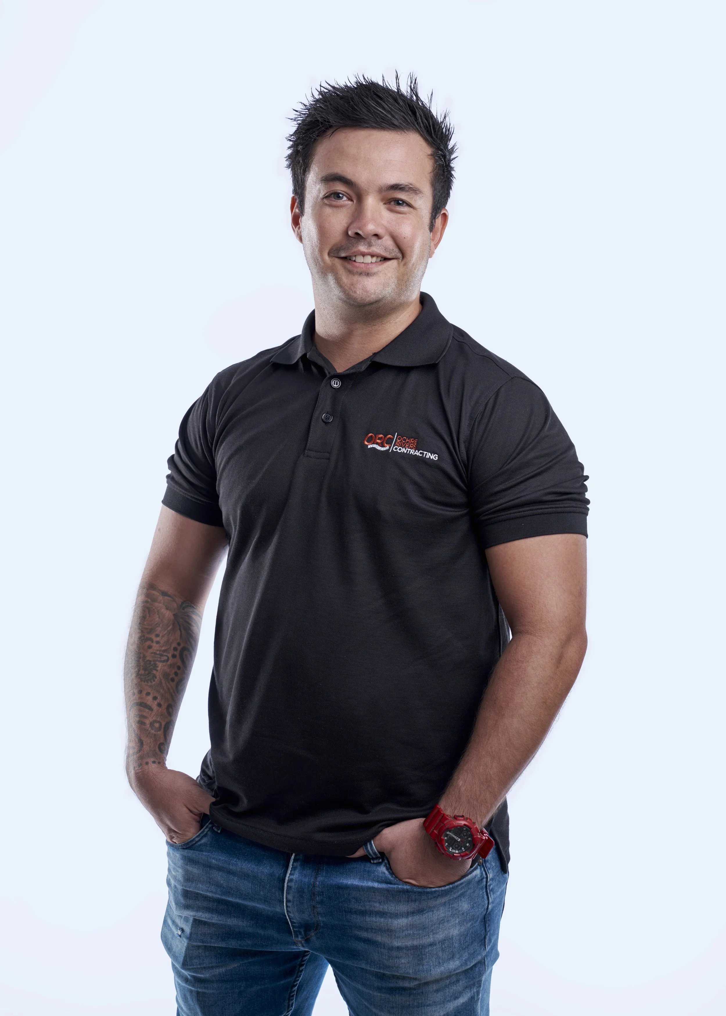 A young man with spiked dark hair, wearing a black polo shirt with a logo, blue jeans, and a red watch, standing with hands in pockets against a light blue background and smiling.