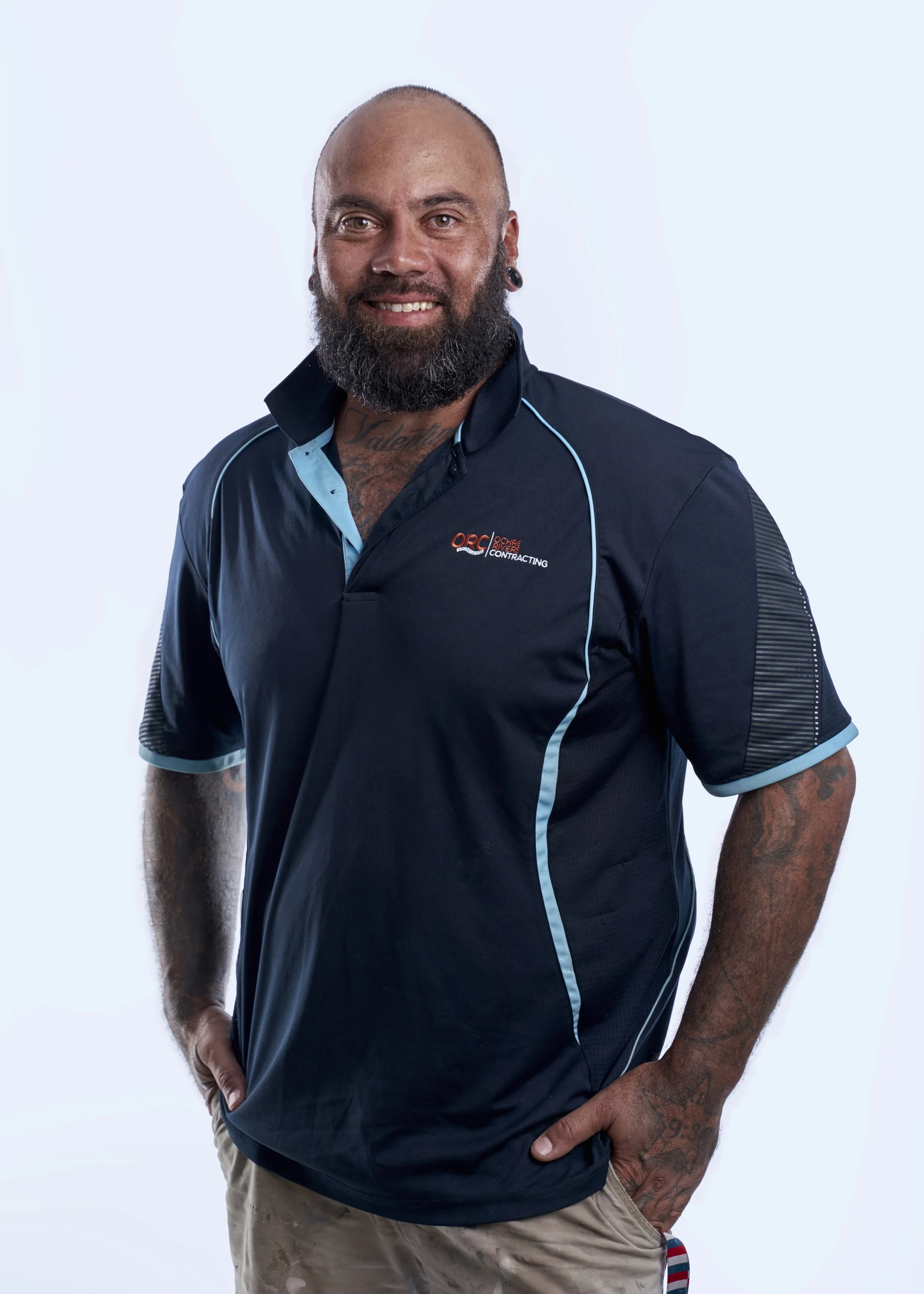 A smiling man with a beard and tattoos wearing a dark blue polo shirt with light blue accents and khaki pants standing against a light background.