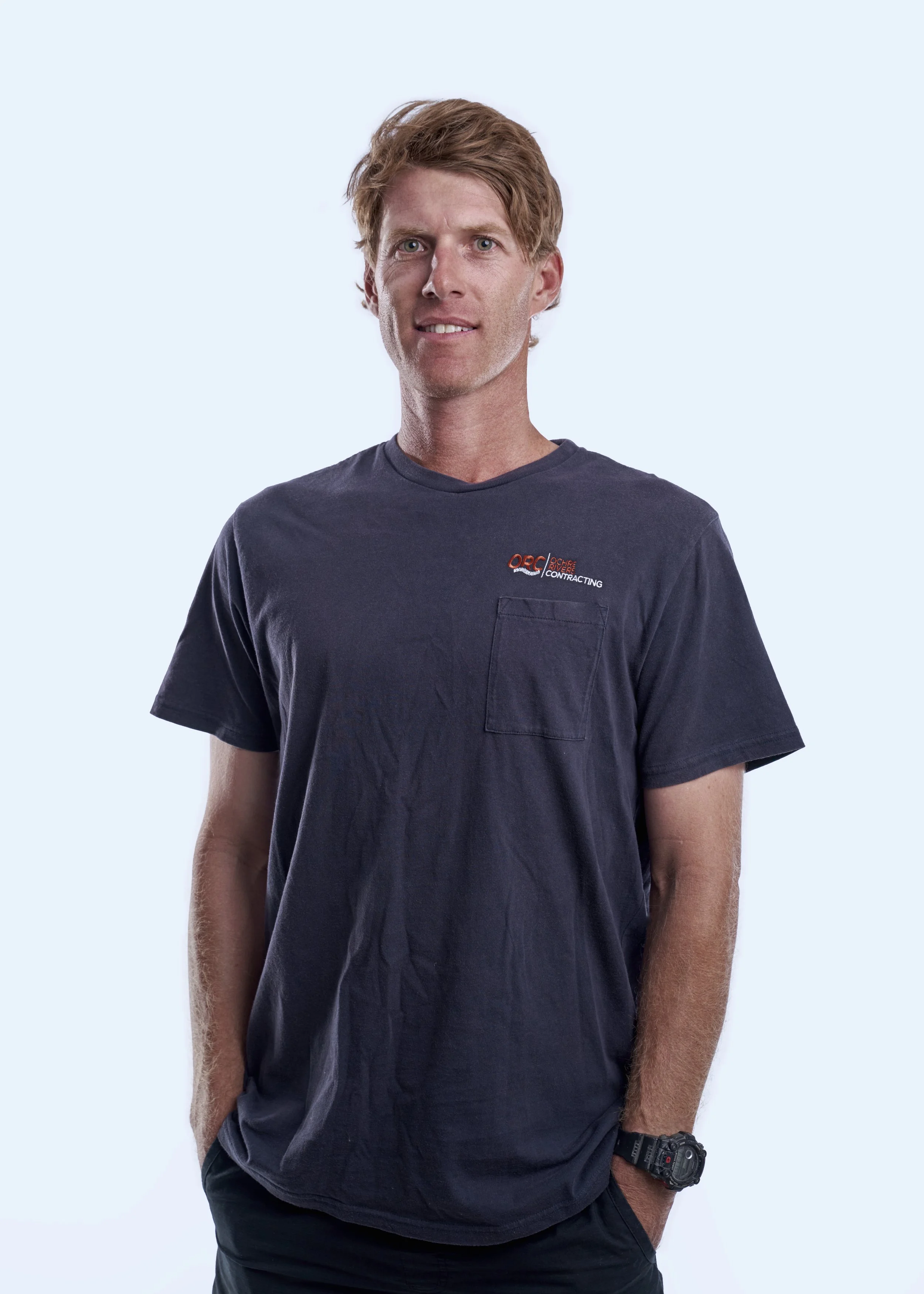 A man with red hair wearing a navy blue T-shirt with a logo on the chest and a black watch on his left wrist. He is standing with his hands in his pockets against a light blue background.