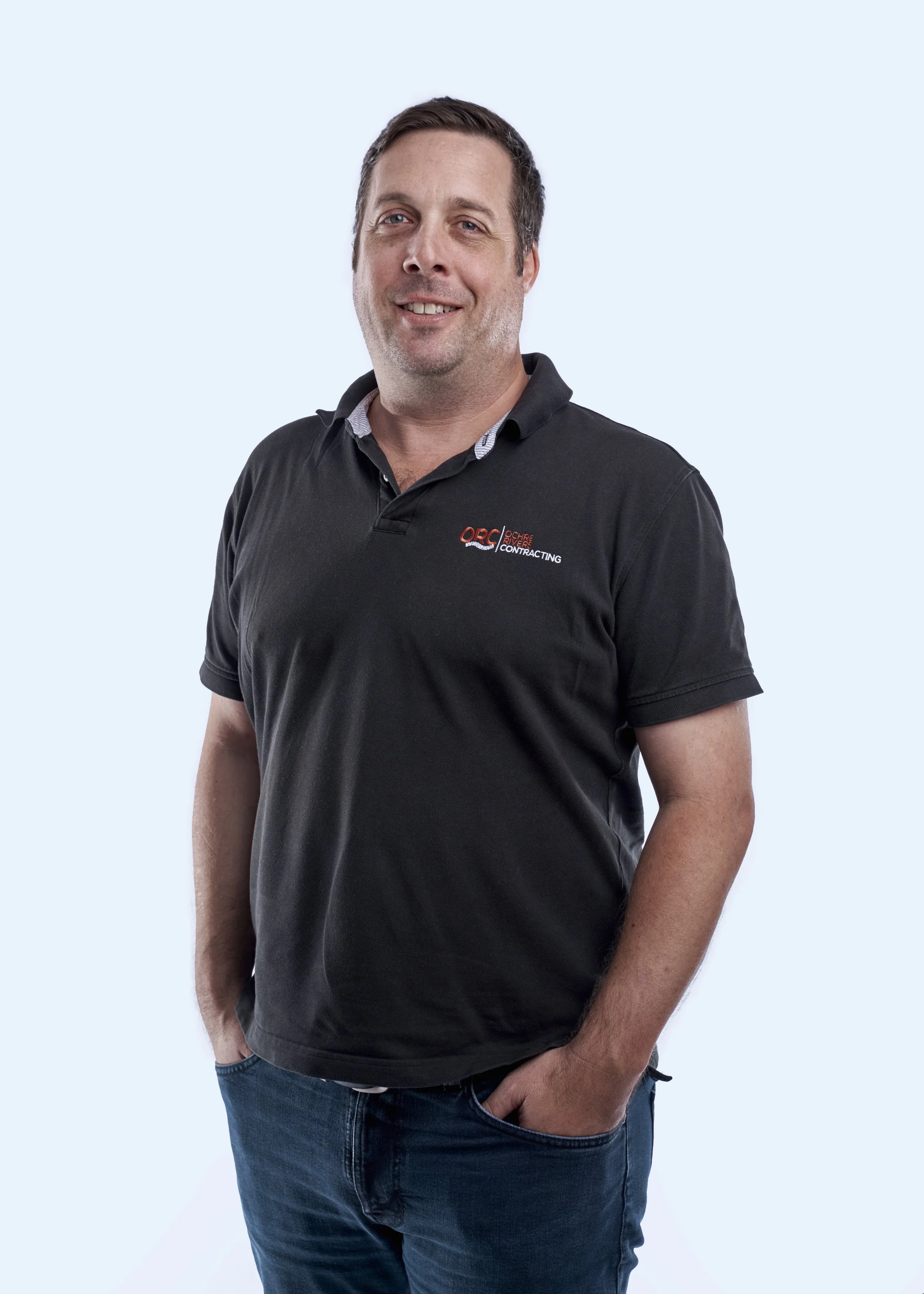 A man smiling and standing with his hand in his pocket, wearing a black polo shirt with a logo, against a light background.