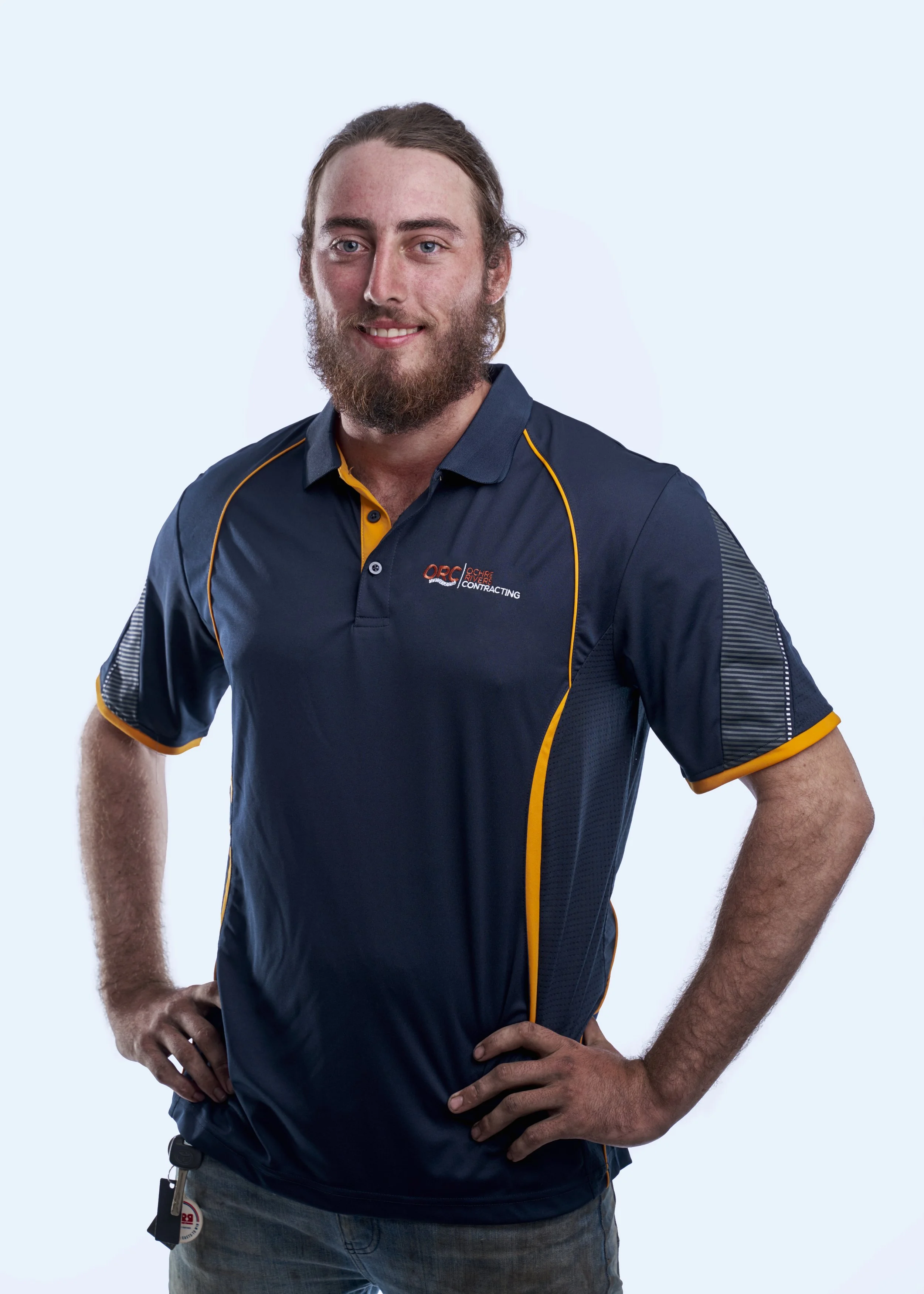 A young man with a beard and long hair, wearing a navy blue polo shirt with yellow accents and the logo 'ORC Contracting,' standing confidently with hands on hips and smiling against a plain light blue background.