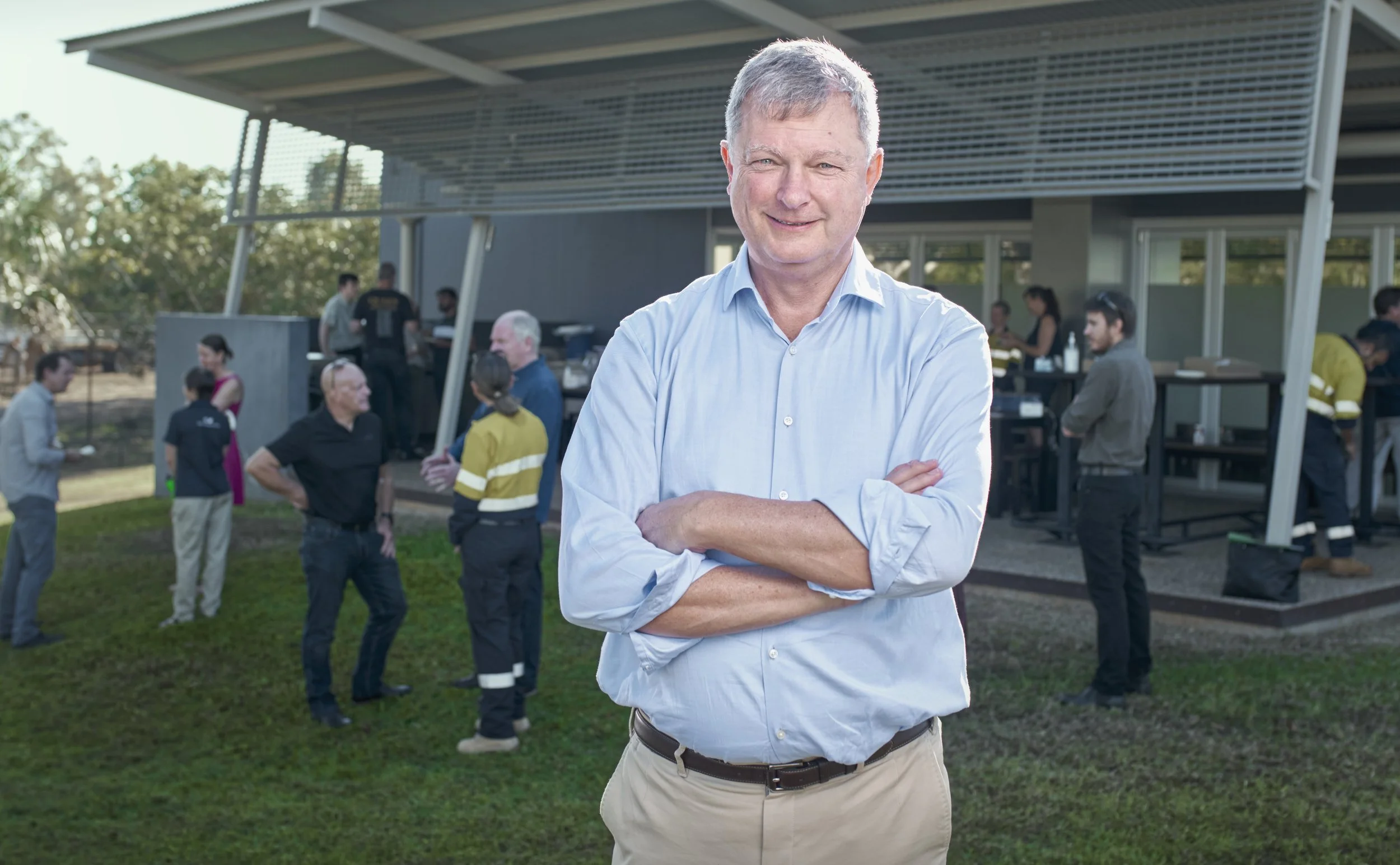 A man in a light blue shirt with arms crossed, smiling, standing outdoors in front of a group of people, including some in uniforms, chatting near a modern building with trees in the background.