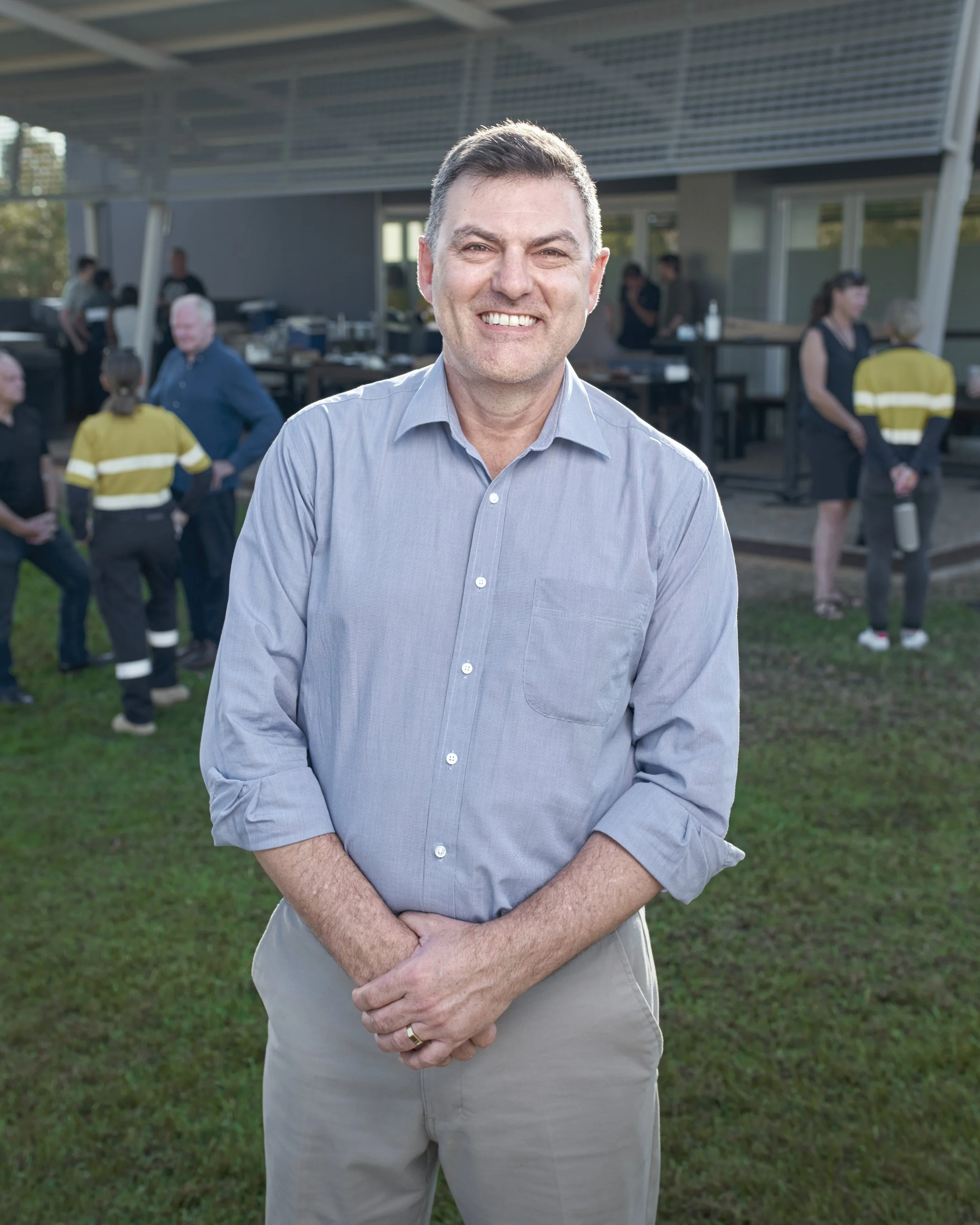 A man in a light gray button-up shirt and khaki pants standing outdoors smiling at the camera, with a group of people, some in yellow reflective safety vests, and a modern building in the background.