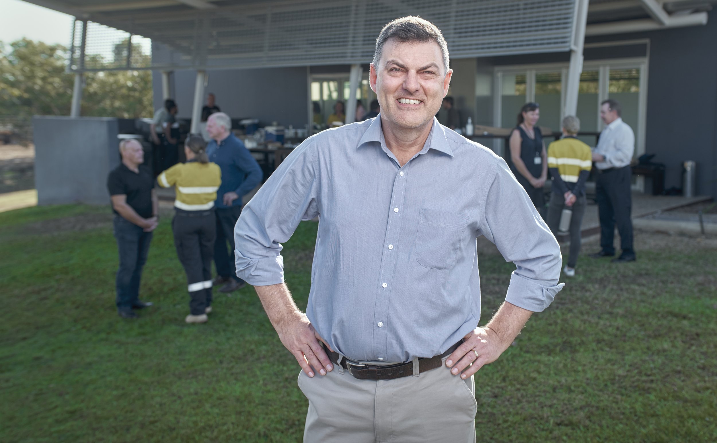 A man in a light blue button-up shirt and khakis standing outdoors with his hands on his hips, smiling at the camera. In the background, several people are gathered, including emergency responders and individuals near a modern building with large win