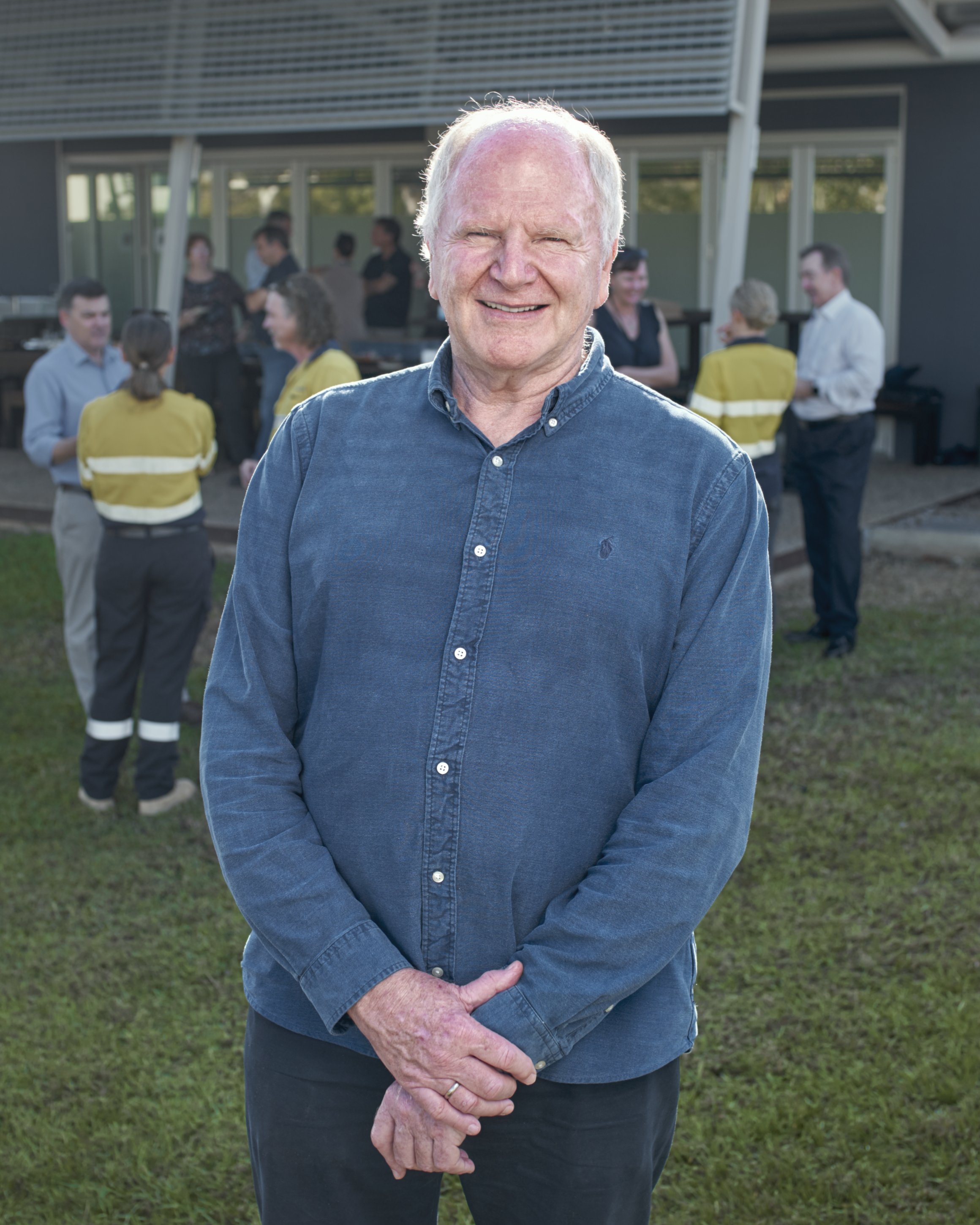 A smiling elderly man in a blue shirt standing outdoors at a gathering with people in the background.