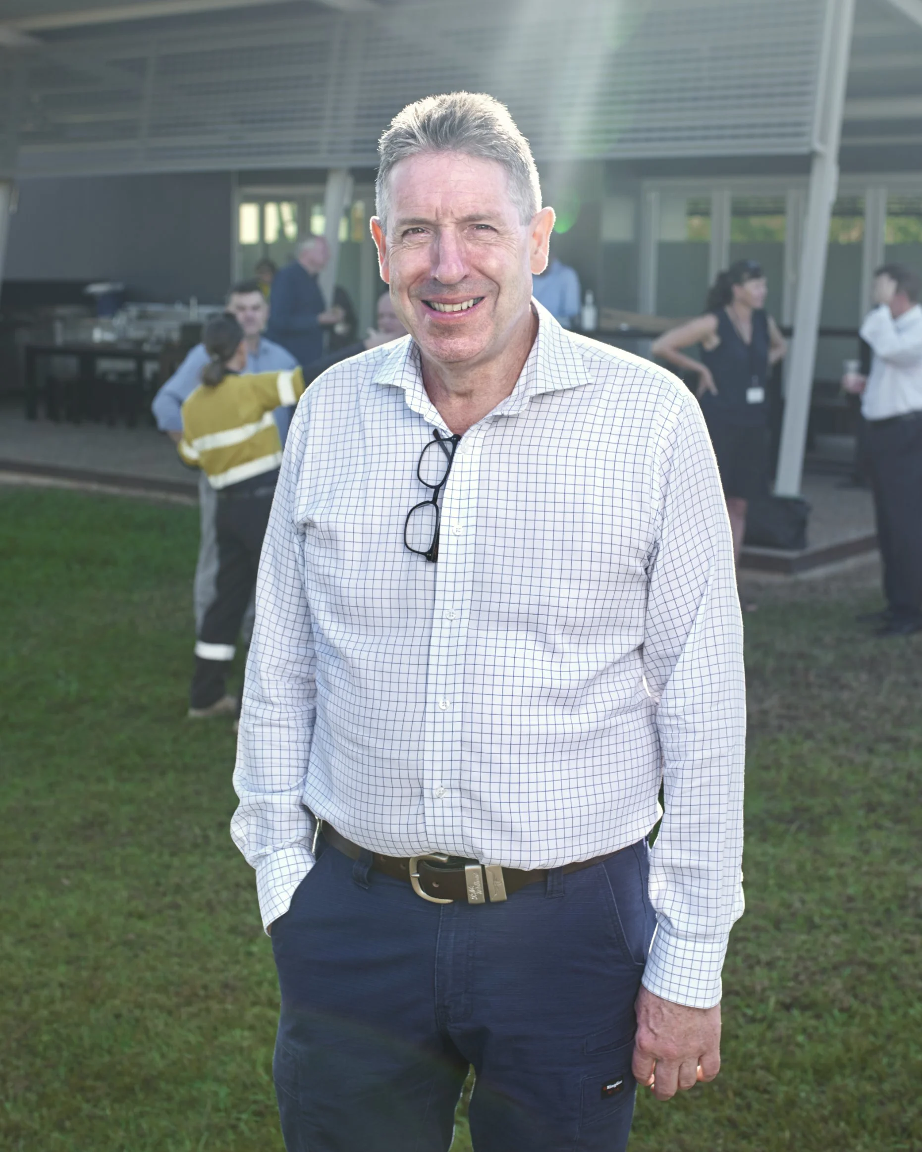 A smiling man with short gray hair wearing a white checkered shirt and dark pants, standing outdoors on green grass during a social event, with people in the background near a modern building.