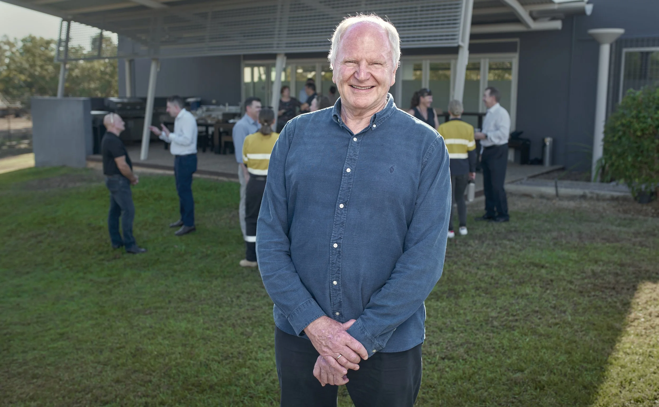An older man with white hair smiling in front of a group of people at an outdoor gathering, with a building and trees in the background.