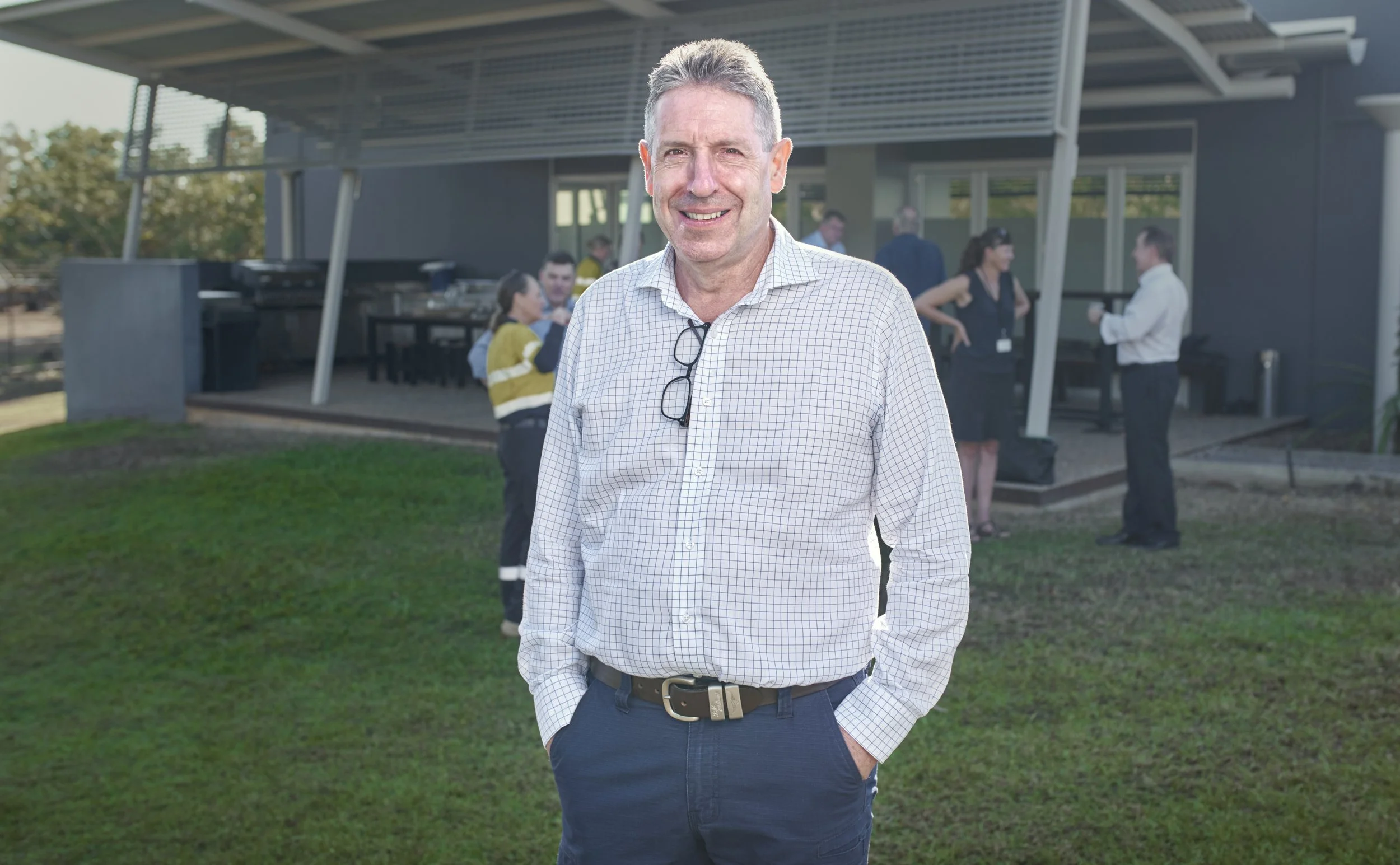 A man in a checkered shirt and blue pants stands outdoors at a social gathering, with a group of people conversing in the background near a modern building.