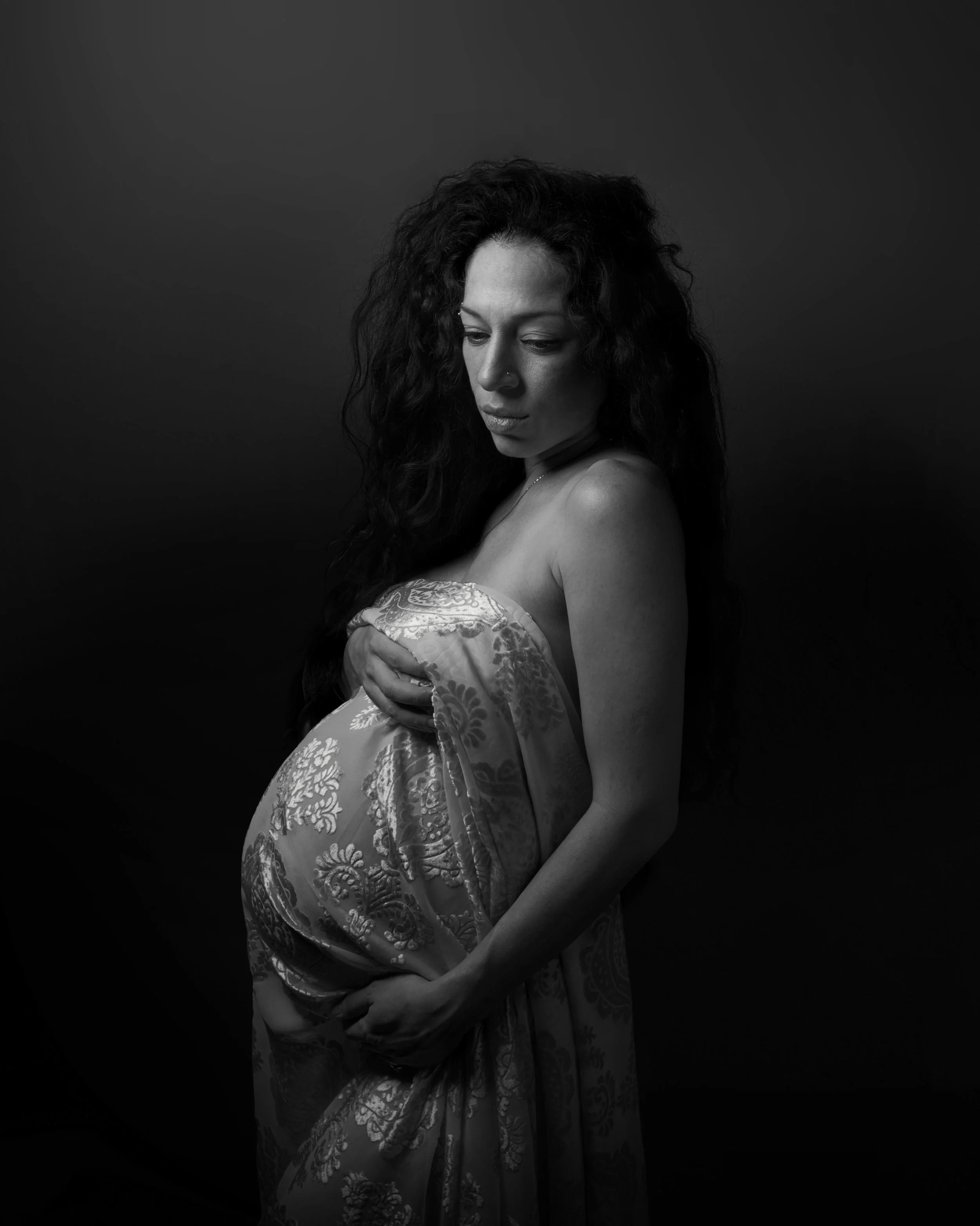 A pregnant woman with curly hair, wrapped in patterned fabric, looking down, photographed in black and white against a dark background.