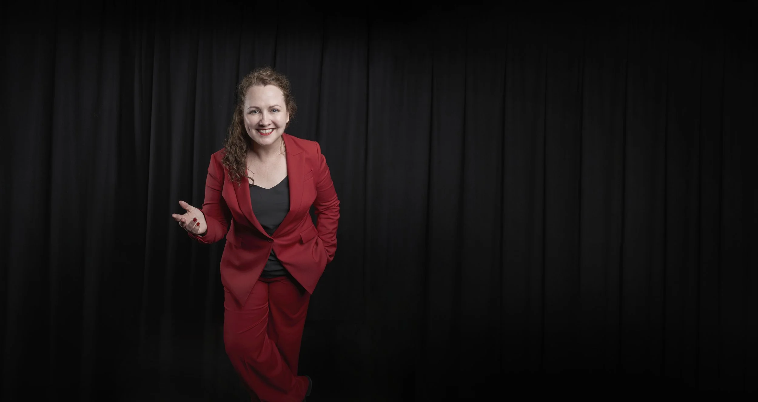 A woman with curly red hair smiling and wearing a red suit standing in front of a black curtain background.