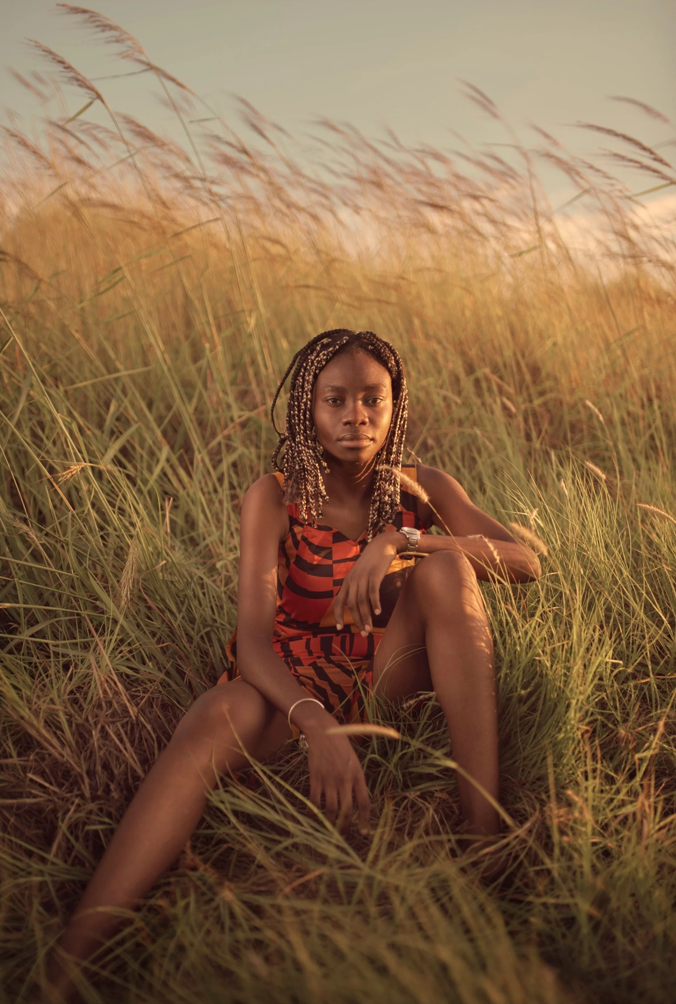A woman sitting in tall grass during golden hour, wearing a patterned dress and sporting braids, with a serious expression.