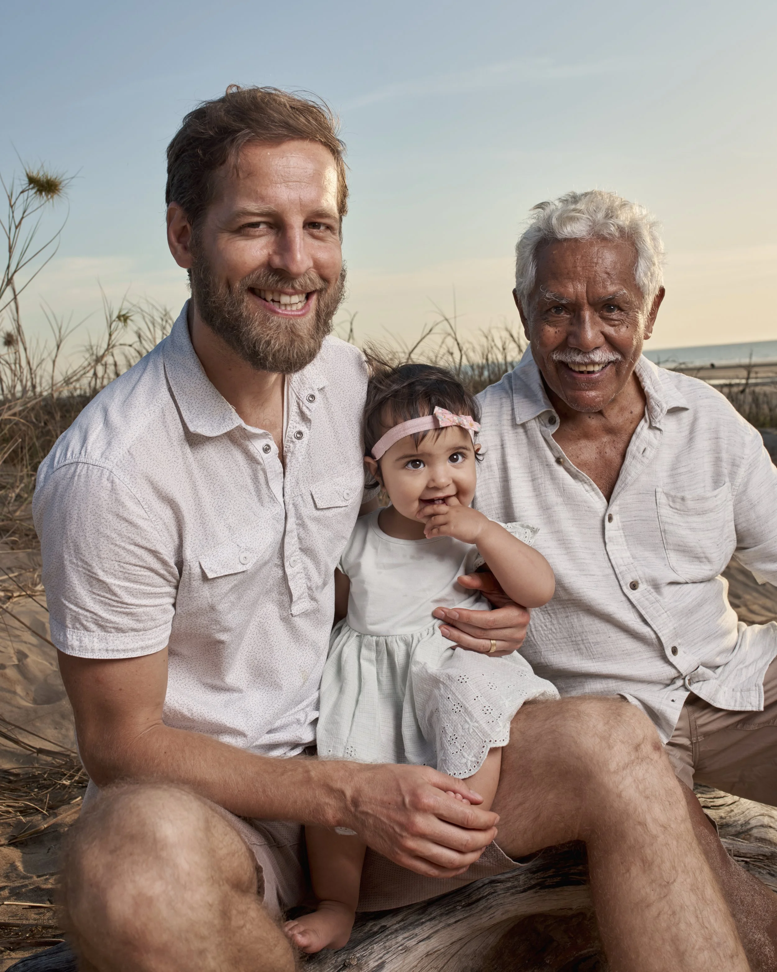 A multi-generational family of three men and a young girl sitting on the beach at sunset, smiling.