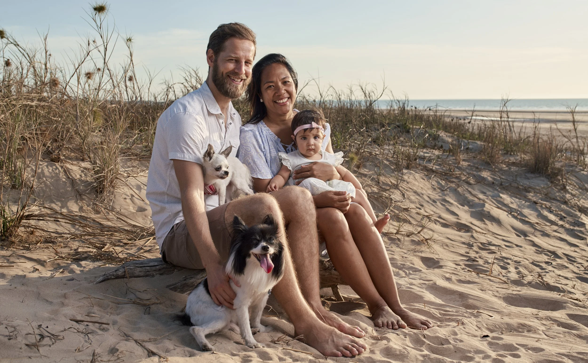 A family of three, a woman, a man, and a young girl sitting on a sandy beach with two small dogs, surrounded by dunes and sparse grass, during sunset.