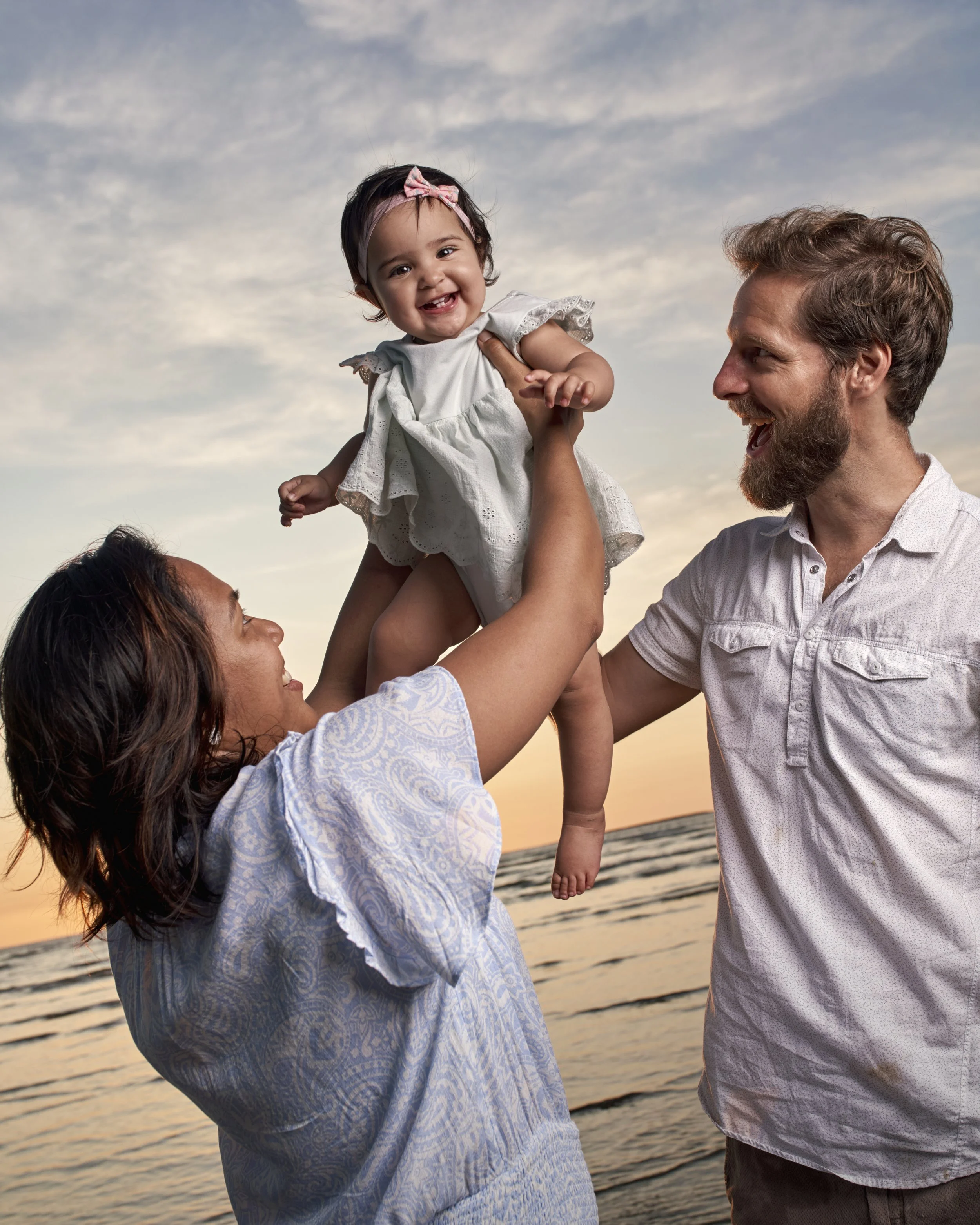 A family of three at the beach during sunset, with a woman lifting a smiling toddler girl, and a man smiling at the girl.