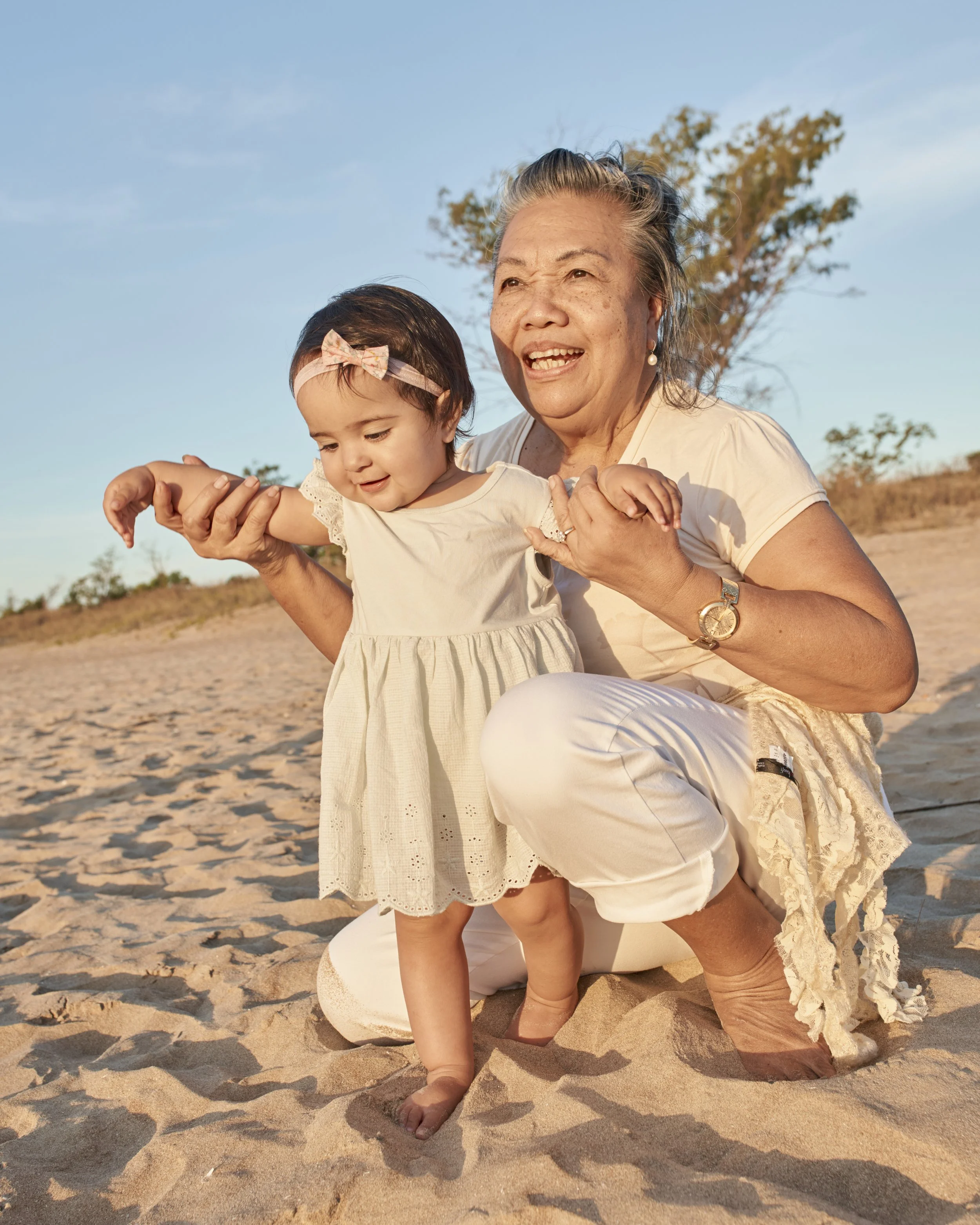 Grandmother and young girl playing on the beach during sunset, holding hands, wearing light-colored dresses, with sand and a tree in the background.