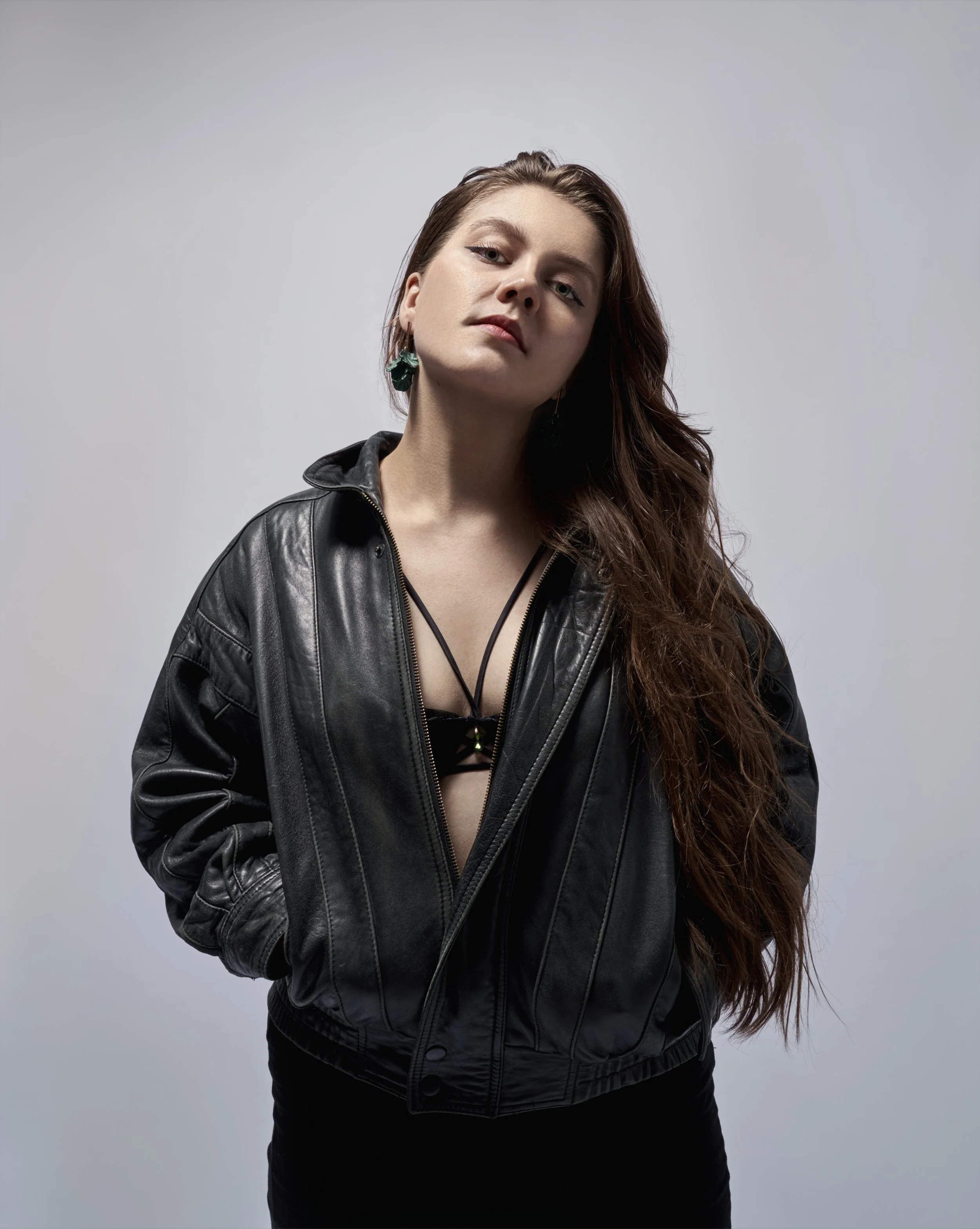 A young woman with long wavy brown hair, wearing a black leather jacket, black top, and green earrings stands against a plain light gray background.