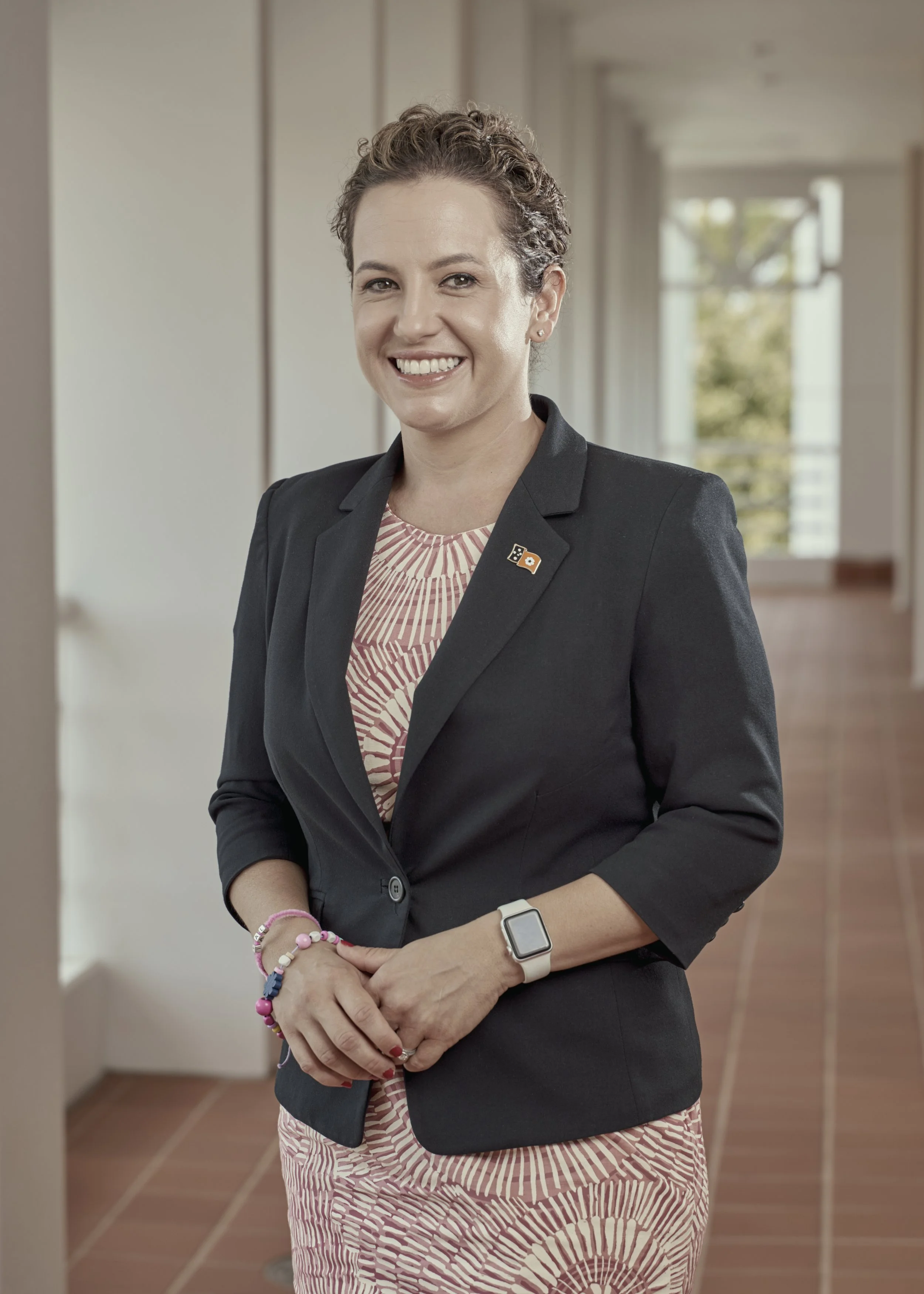 A smiling woman in a professional black blazer and patterned dress standing in a well-lit indoor corridor, wearing a watch and colorful bracelets.