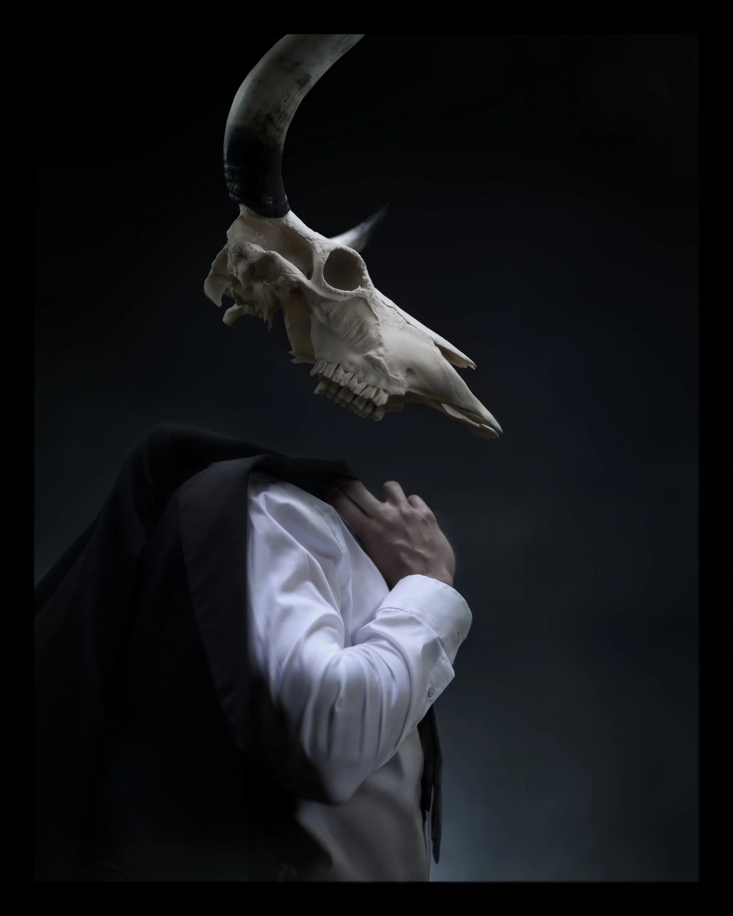 Person in white shirt and black jacket with hand on chin, wearing a horned animal skull mask, against dark background.