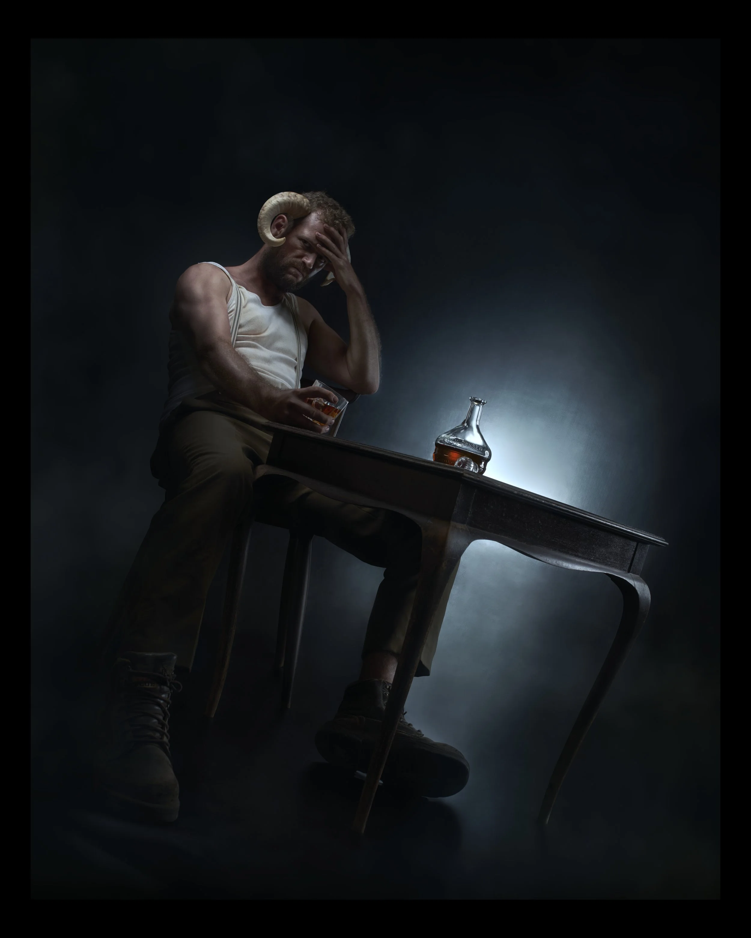 A man with ram horns, wearing a white tank top and jeans, sitting at a wooden table with a bottle of alcohol and a glass, holds his head in frustration in a dimly lit setting.