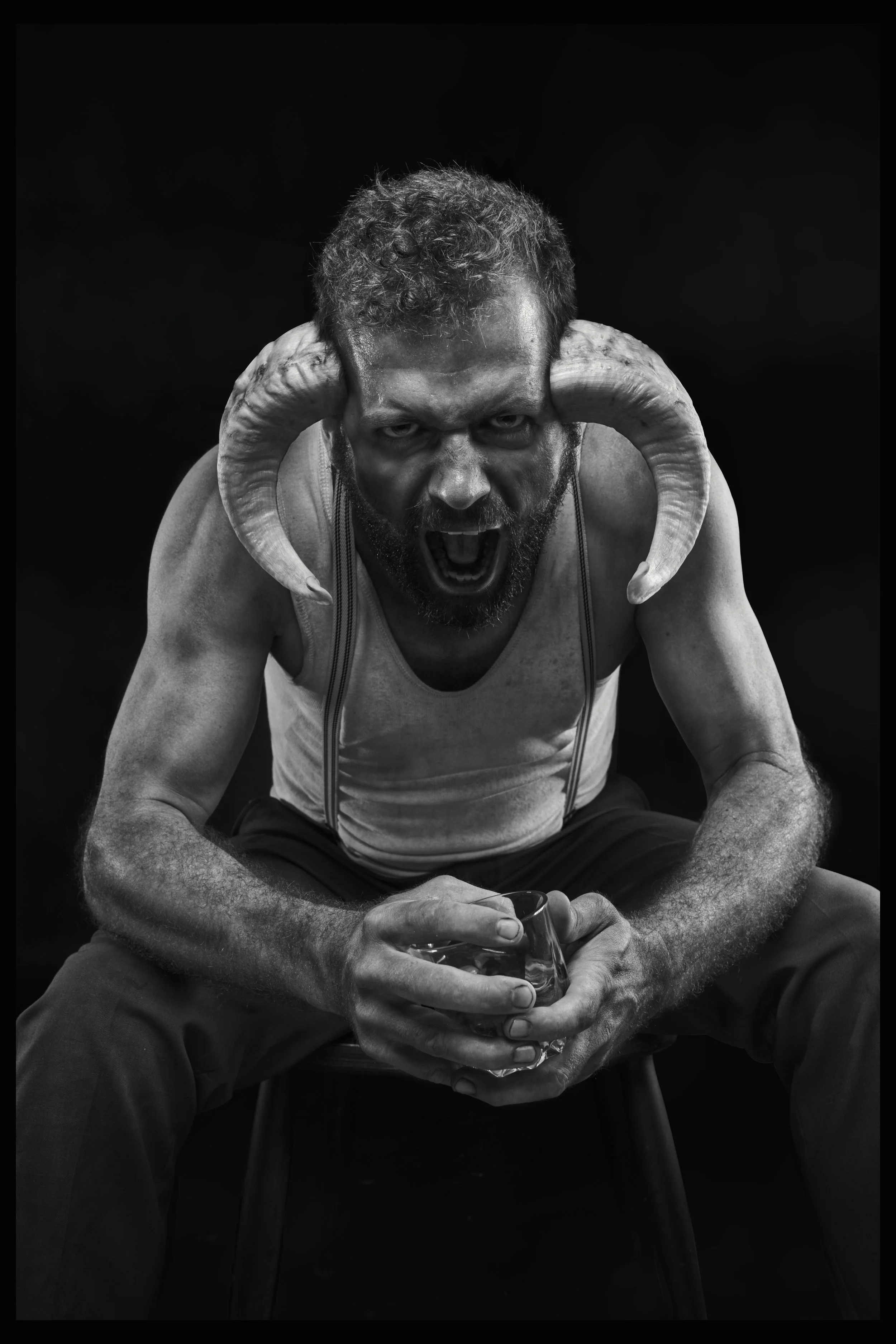 A black and white photo of a man with a beard and curly hair wearing a tank top, with ram horns on his head, sitting and yelling while holding a glass of water.