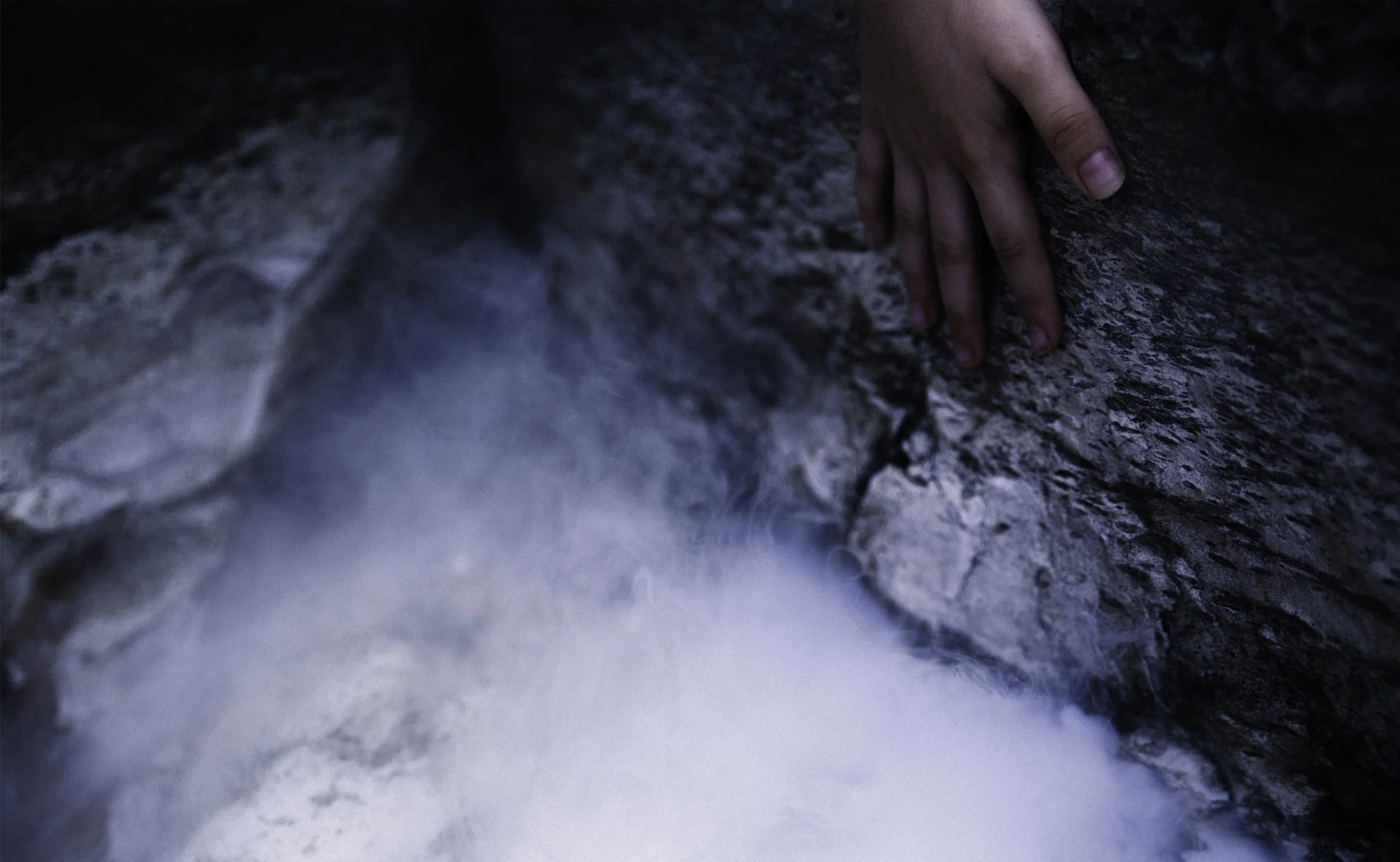 A person's hand leaning on a rough rock formation near rushing water.