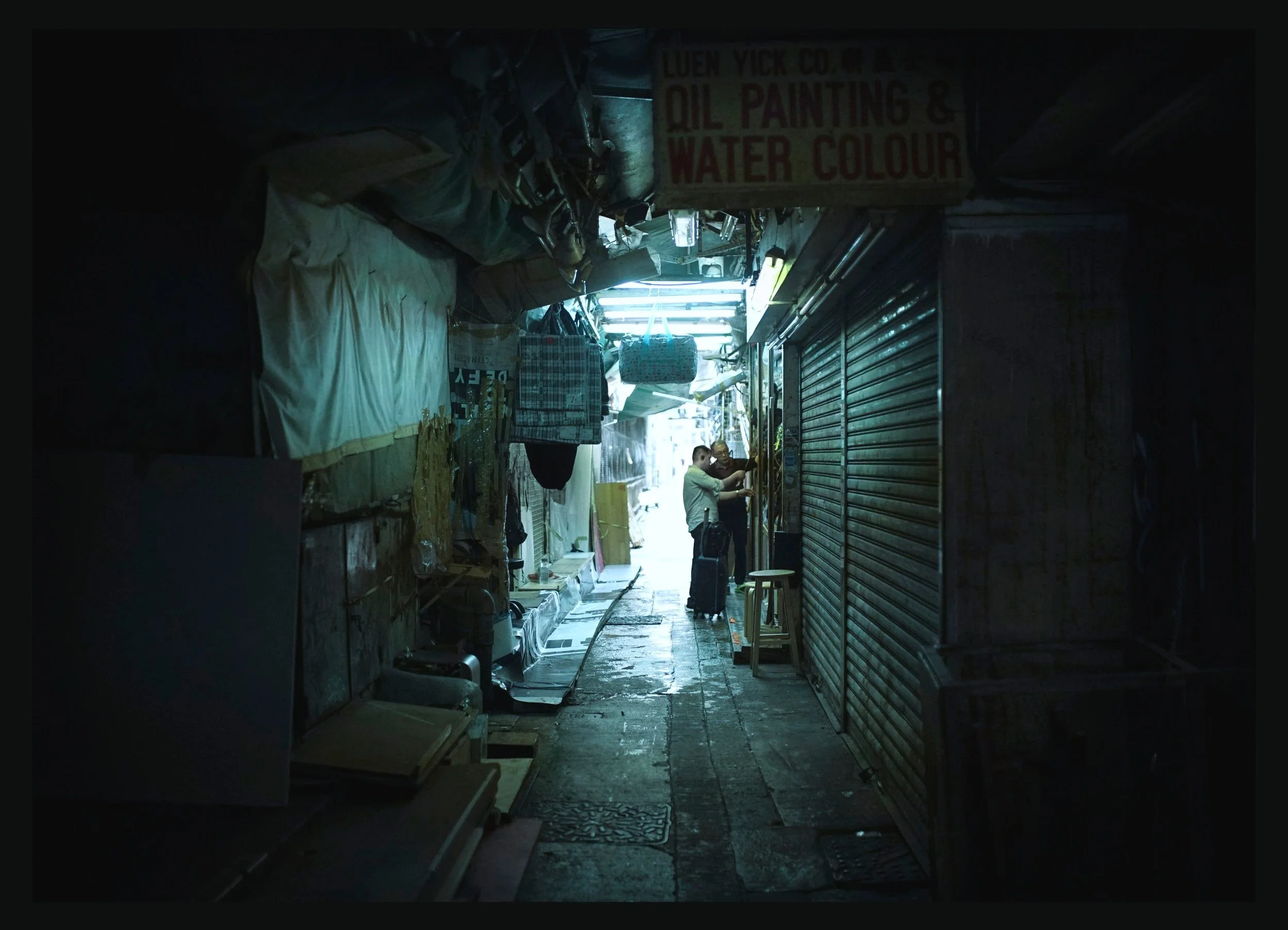 A narrow alleyway market with closed metal shutters on the right and hanging bags and fabrics on the left. Two people are standing near the center, possibly adjusting a display or sign. Bright light at the end of the alley contrasts with the darker f