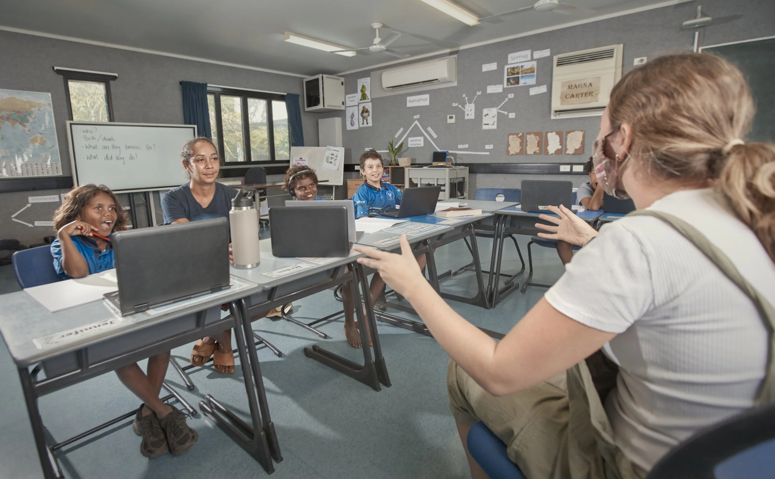 Teacher wearing face mask and glasses speaking to a classroom of young students seated at desks with laptops and notebooks in a classroom with educational posters and whiteboard.