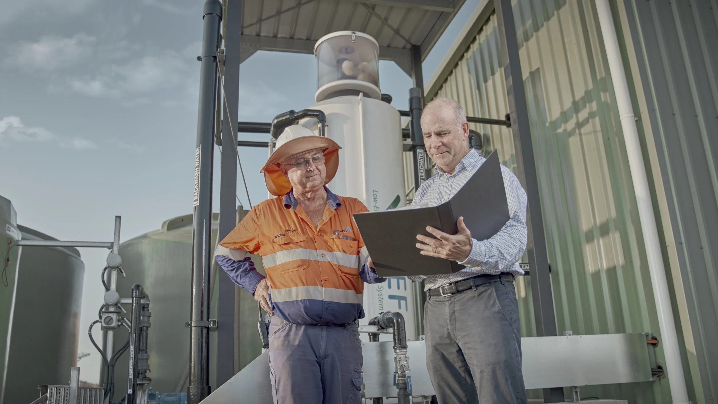 Two men, one in an orange safety shirt and wide-brimmed hat, and another in a light dress shirt, are outside near some industrial equipment, examining a clipboard together.