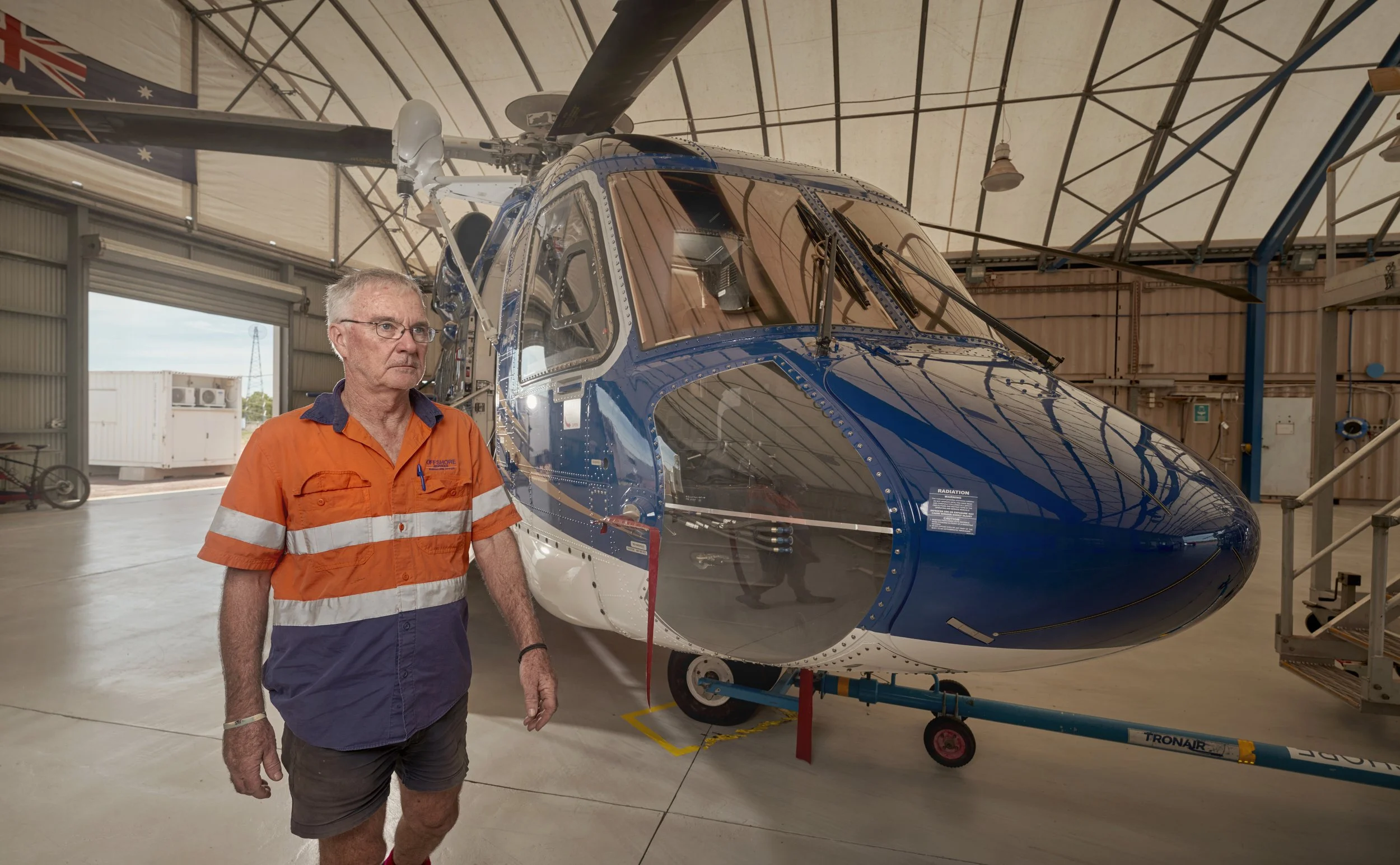 An older man in an orange and blue shirt walking in an aircraft hangar next to a blue helicopter.