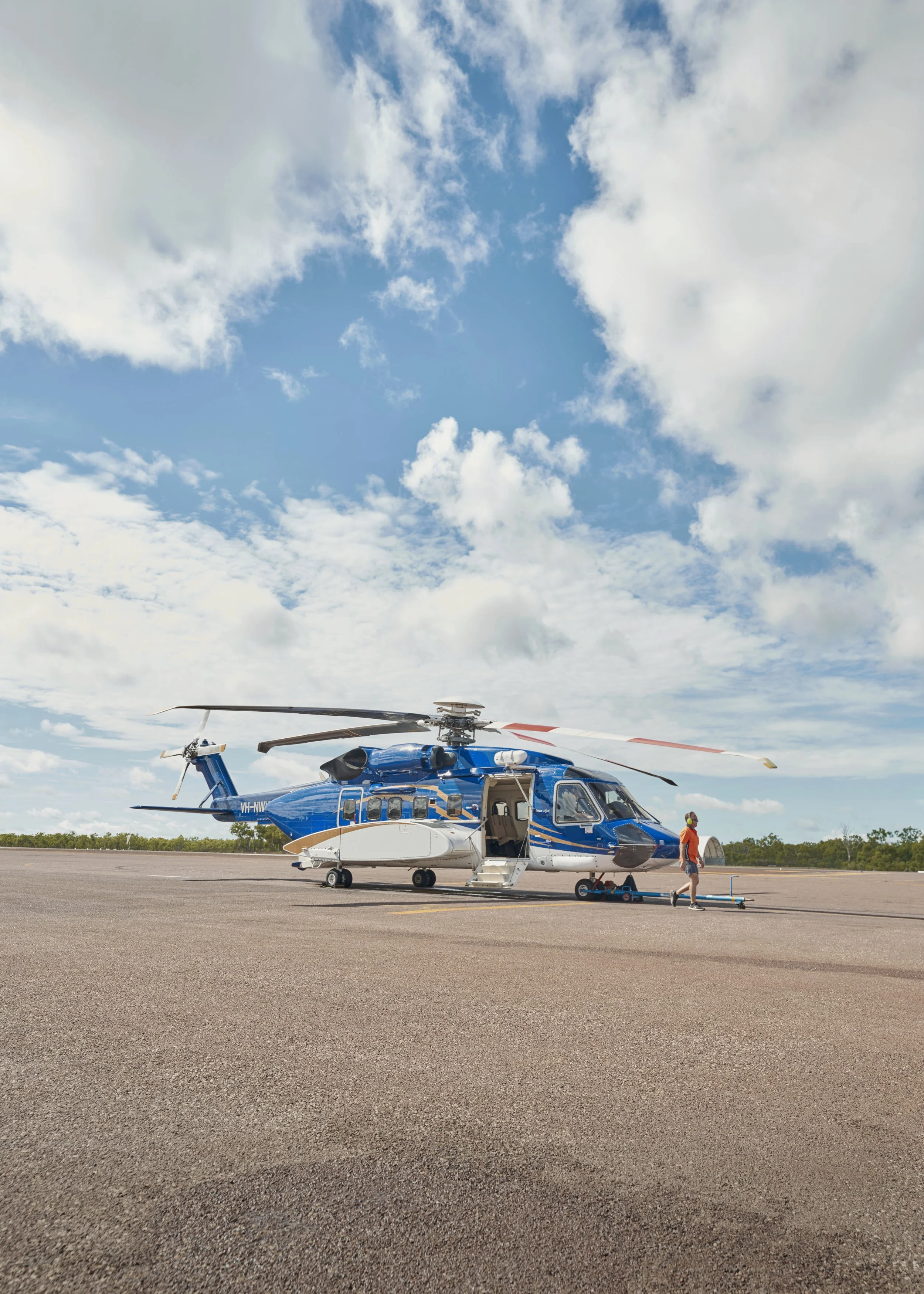 Blue and white helicopter parked on a tarmac with an open door and a person walking nearby, under a partly cloudy sky.